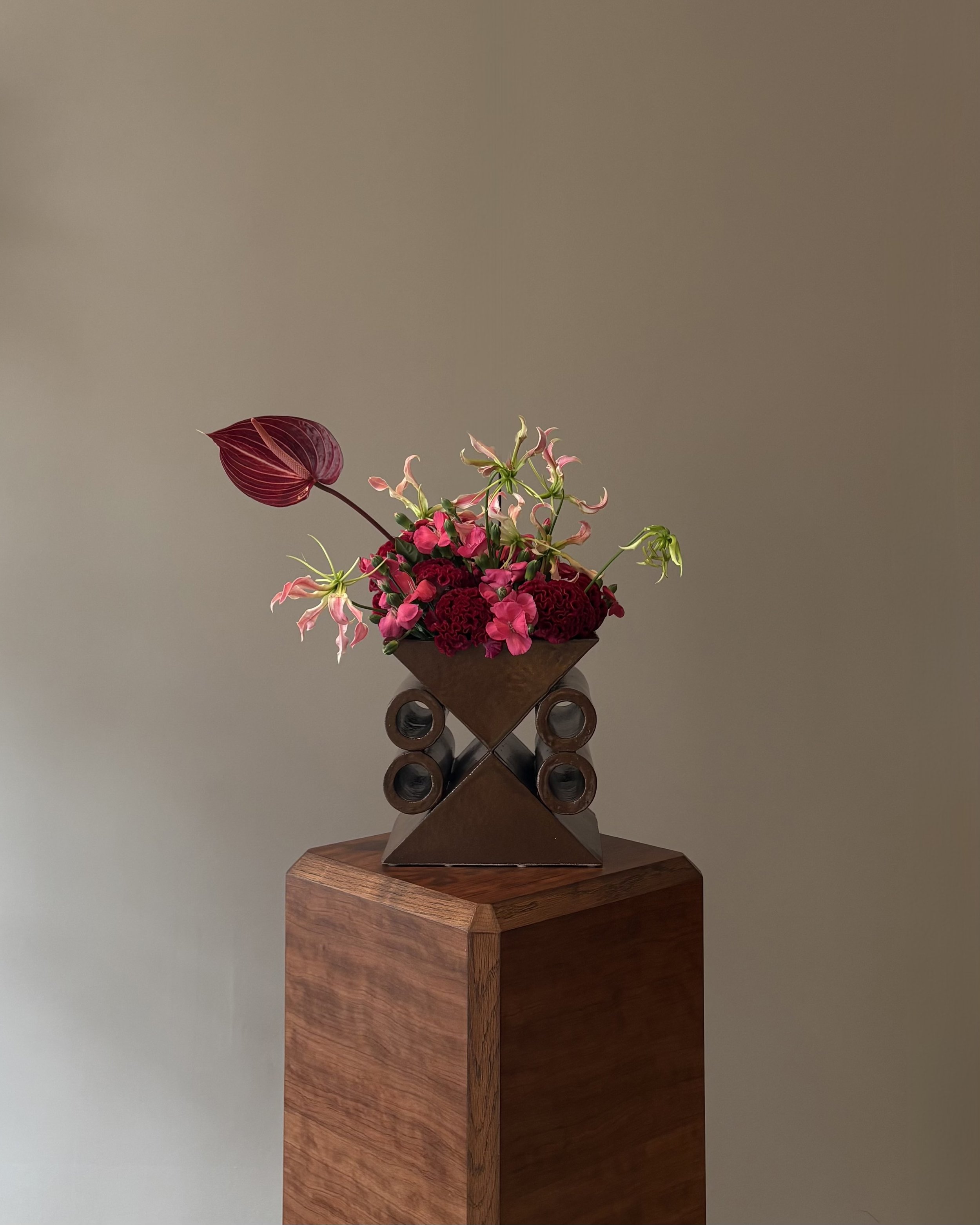 A bouquet of pink and red flowers in a modern, geometric vase on a wooden pedestal against a plain, neutral background.