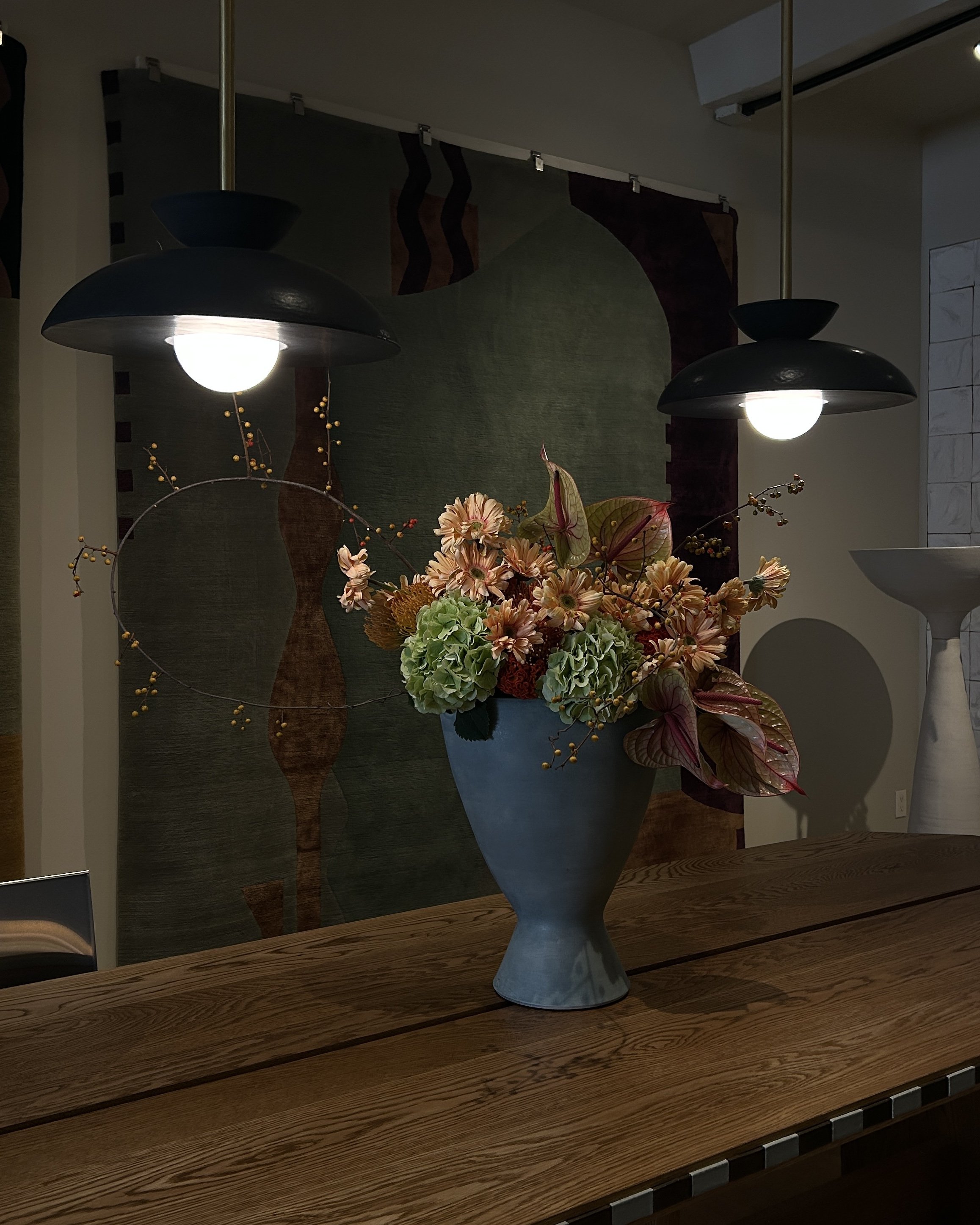 A blue vase filled with pink, orange, and green flowers on a wooden table, illuminated by two overhead lights in a room with abstract wall art.