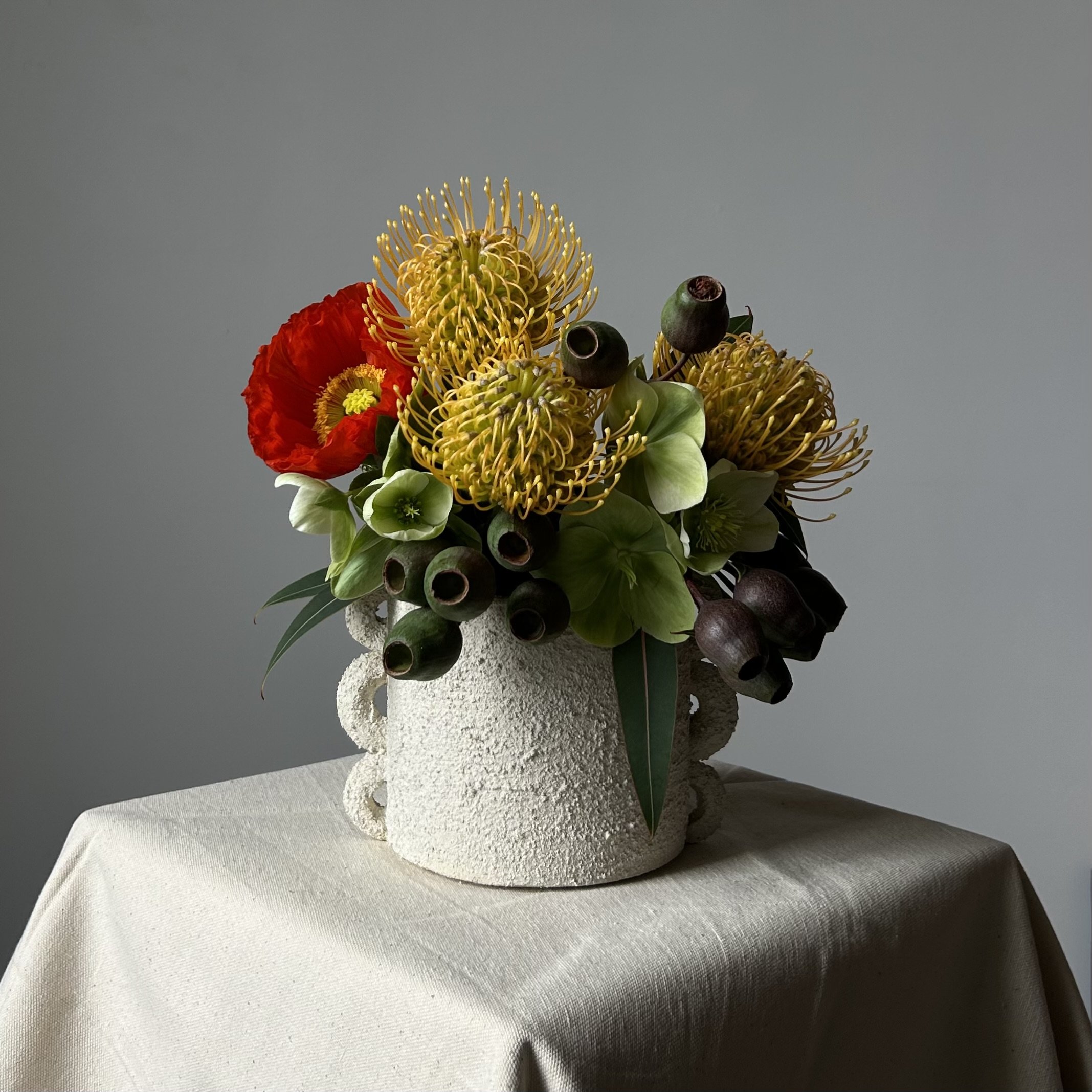 A textured white ceramic vase with an arrangement of colorful flowers, including yellow pincushion proteas, red poppies, green hellebores, and dark seed pods, on a table covered with a cream-colored cloth.
