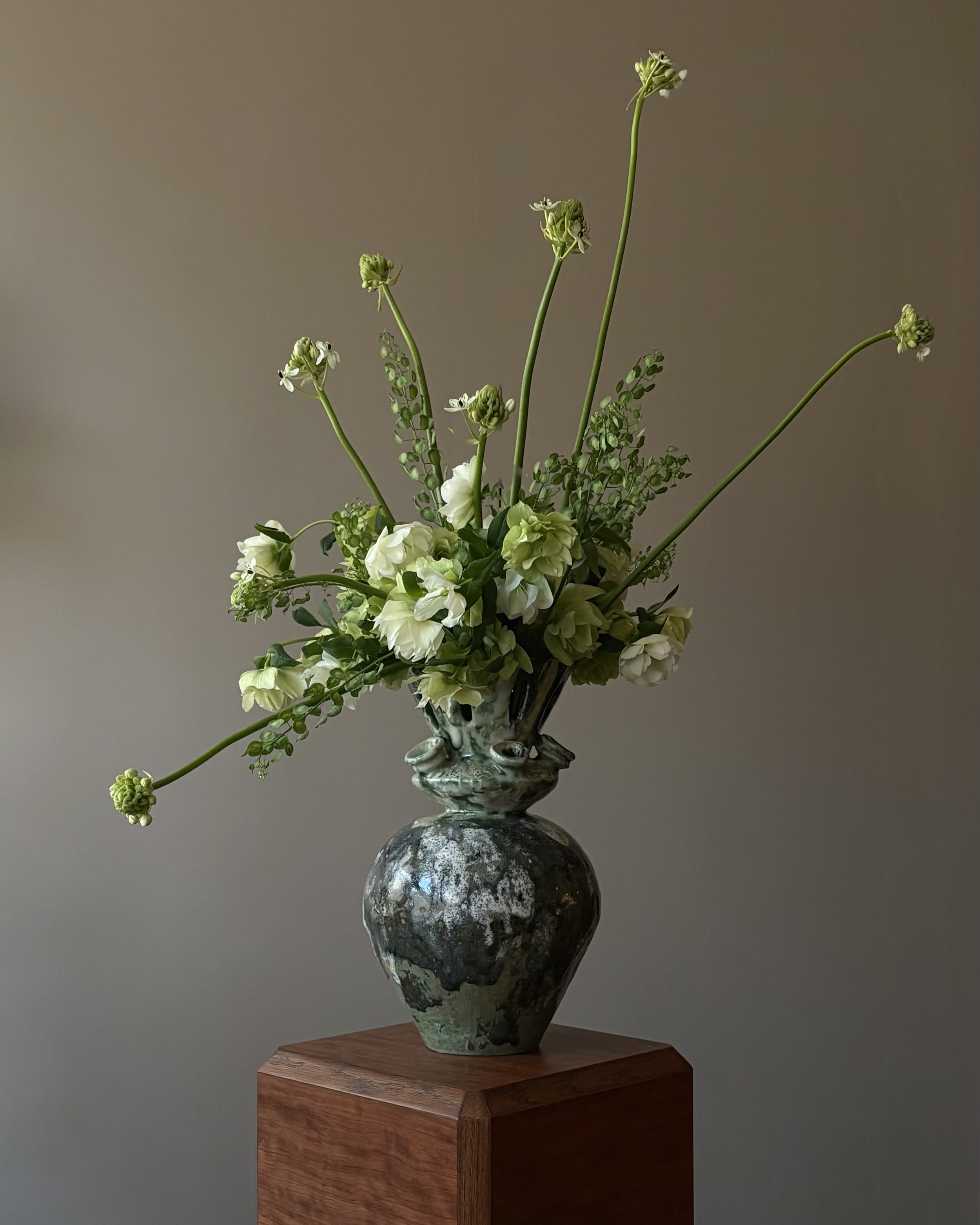 A ceramic vase with a green and white marbled design holding white and cream-colored flowers and green foliage, placed on a wooden pedestal.