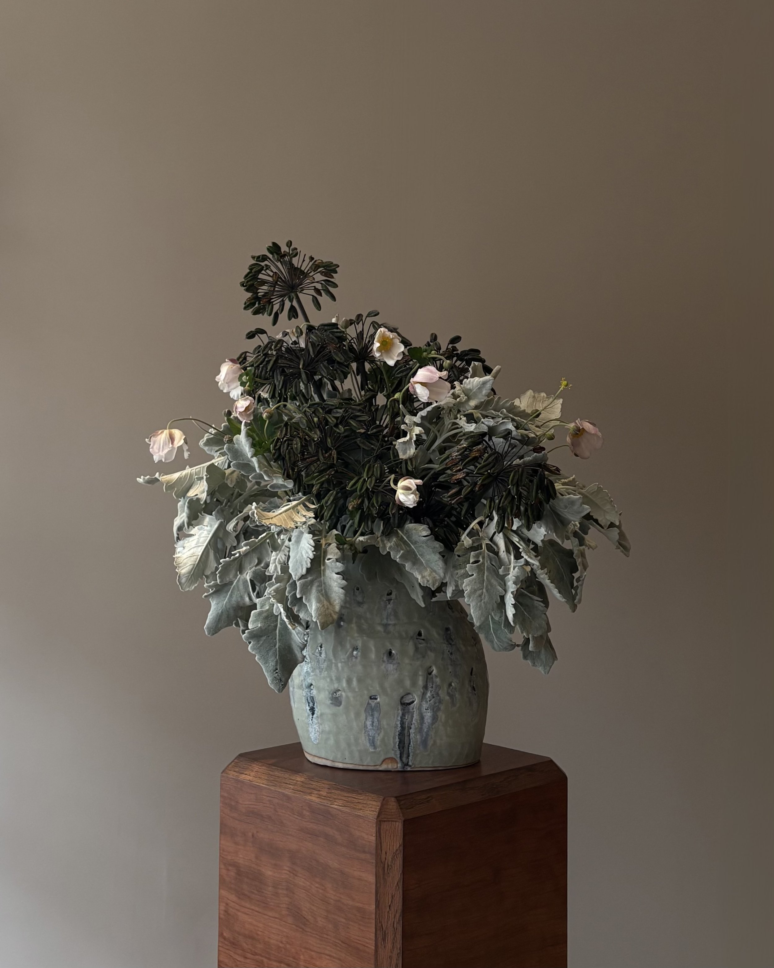 A ceramic vase with dried and fresh flowers, including white and pink blossoms and large gray-green leaves, sitting on a wooden pedestal against a plain beige wall.