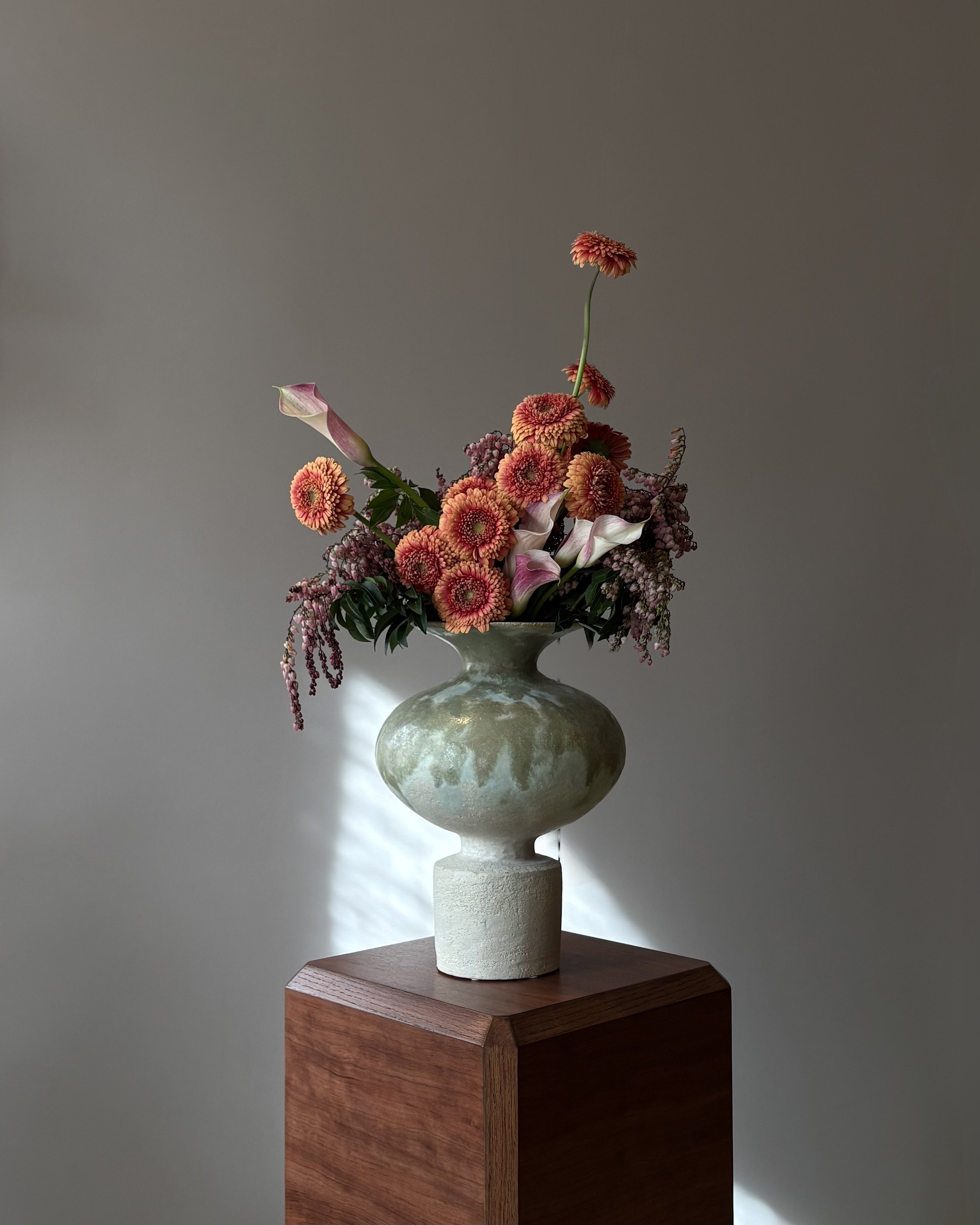 A flower arrangement in a green ceramic vase, placed on a wooden pedestal, with pink, purple, and orange flowers and greenery.