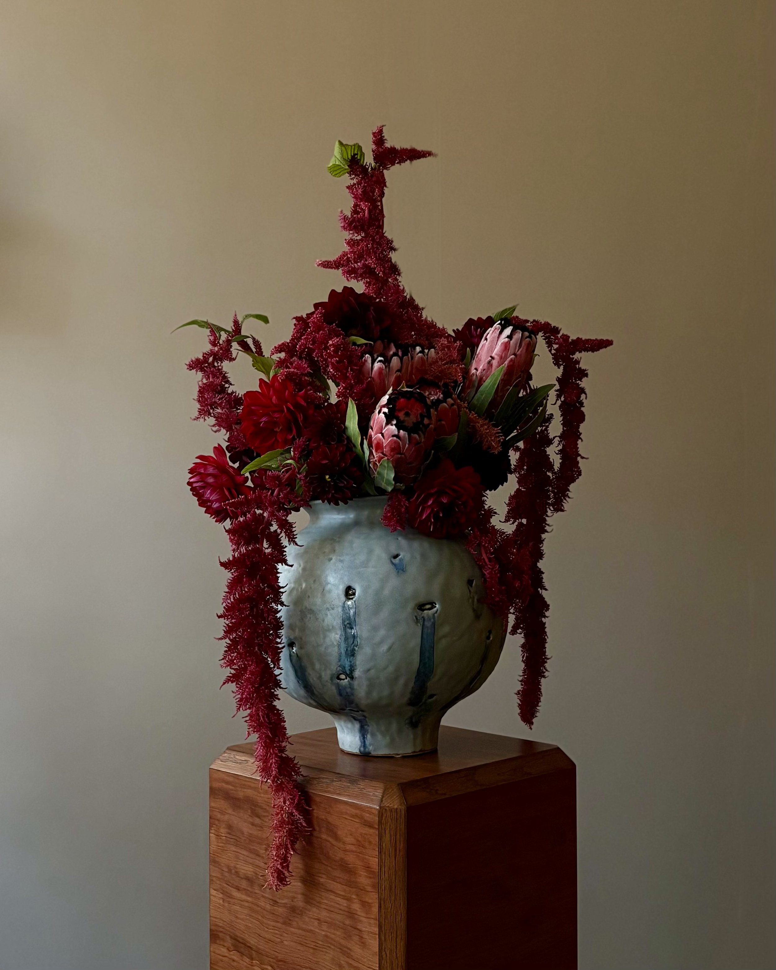 Ceramic vase with textured glaze holding a bouquet of red and pink flowers, including proteas and other blooms, on a wooden pedestal against a plain wall.