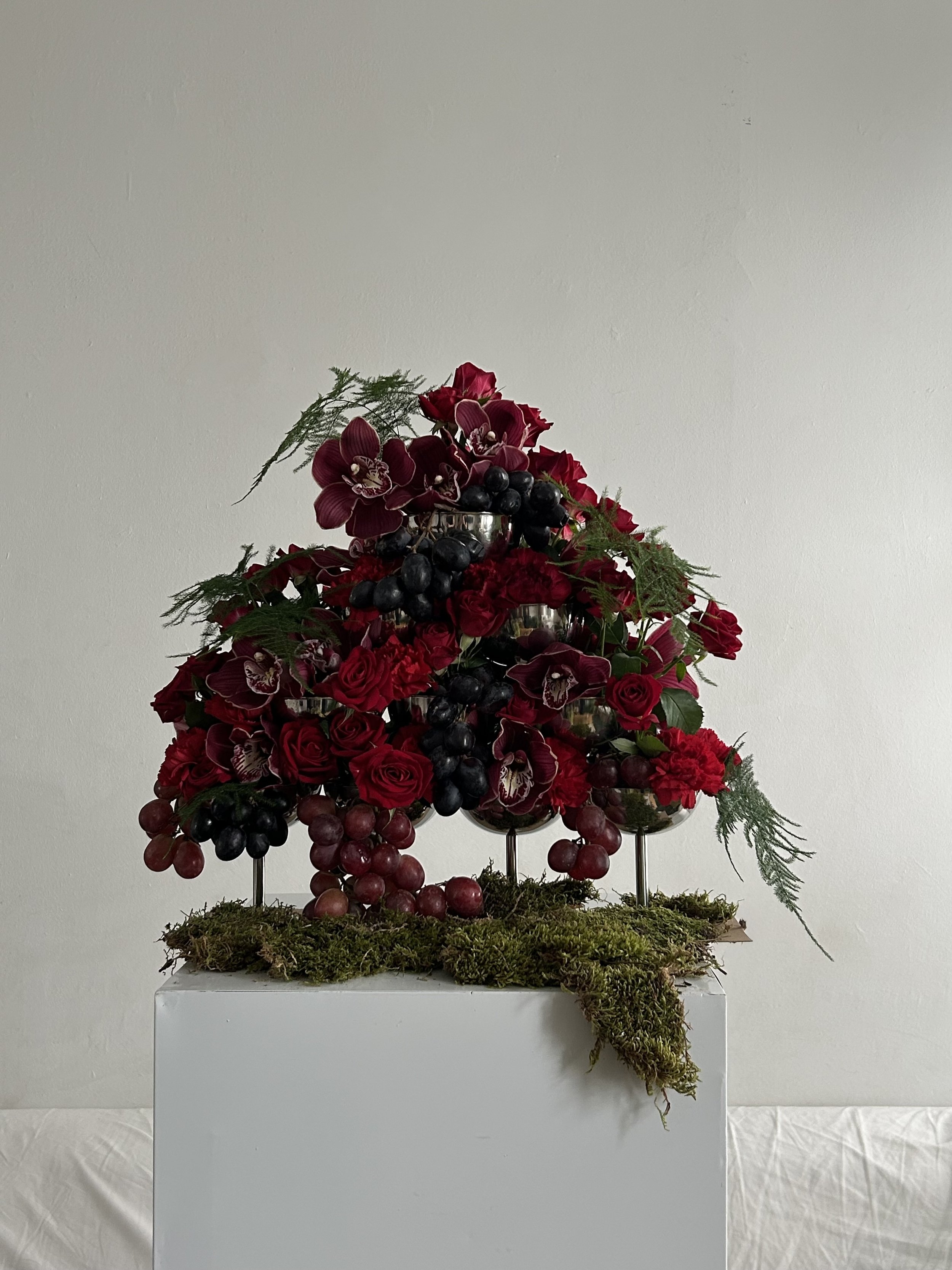 Decorative floral arrangement with red roses, orchids, grapes, and greenery on a white pedestal against a plain wall.