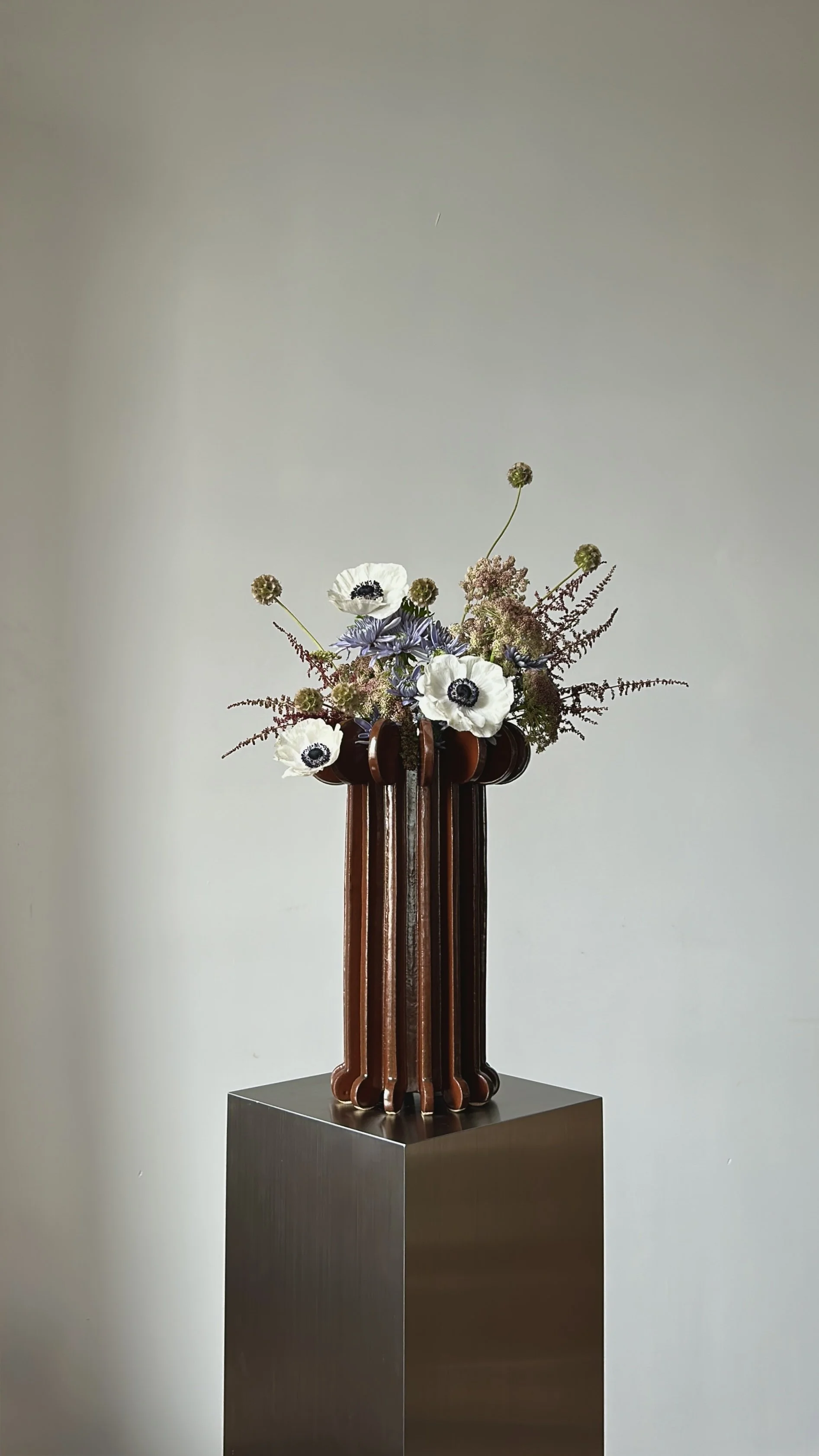 A floral arrangement with white anemones and purple flowers in a tall, wooden vase on a metallic pedestal.