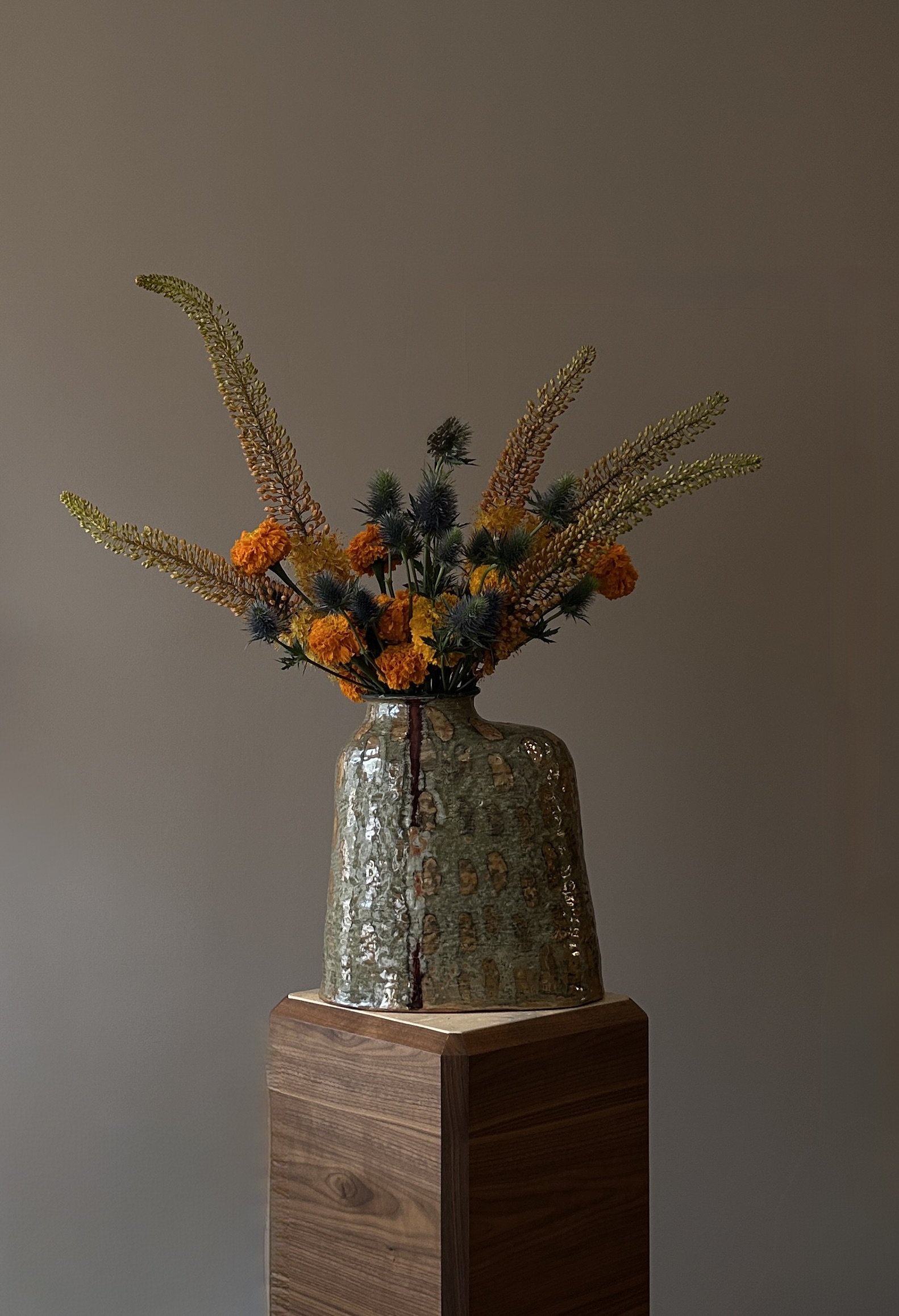 A ceramic vase with a textured, reflective surface, filled with a mixed arrangement of dried flowers and plants, sitting on a wooden pedestal against a plain wall.