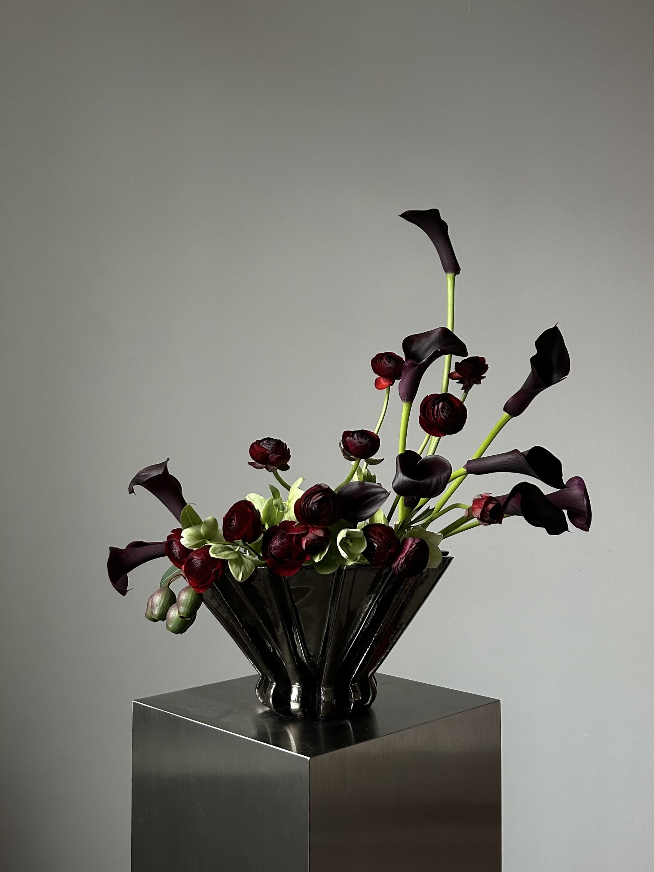 A modern black metallic vase with dark purple calla lilies and red ranunculus flowers, arranged on a metallic pedestal against a plain gray background.