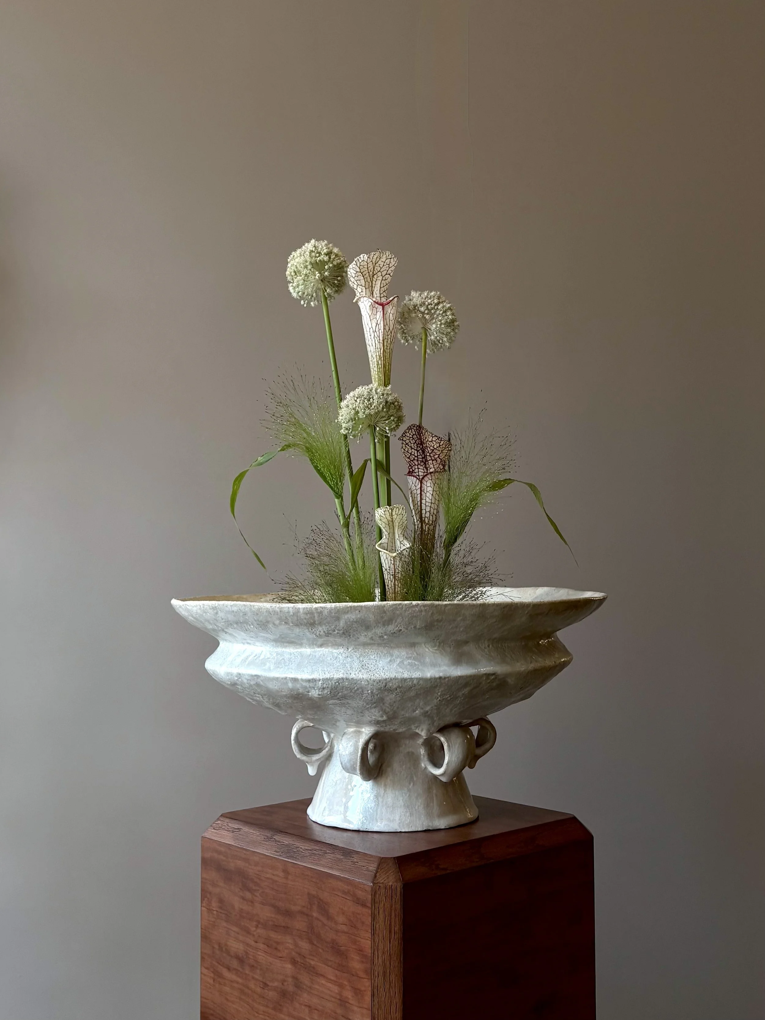 An artistic floral arrangement in a white, textured ceramic vase on a wooden pedestal, featuring various white and green flowers and leaves.