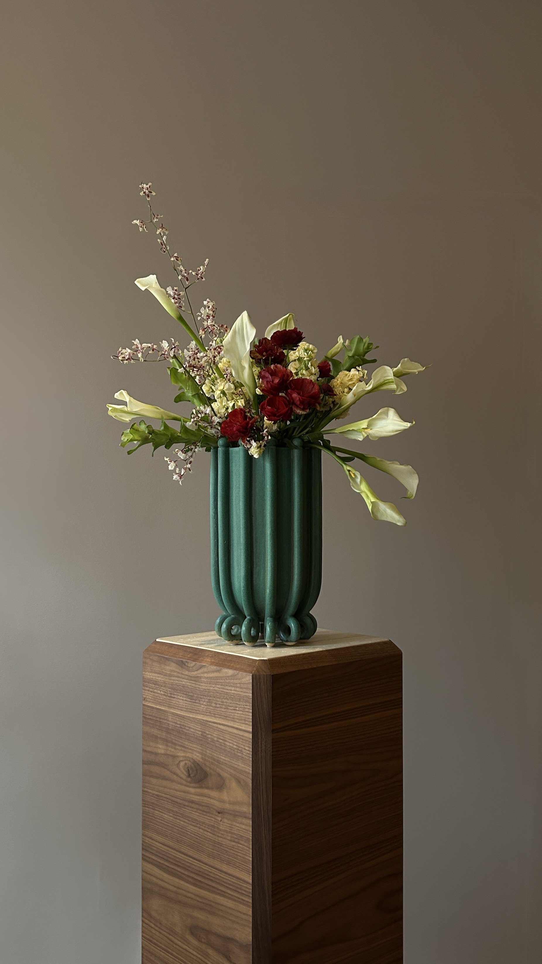 A flower arrangement in a tall, green vase with vertical ridges sits on a wooden pedestal.