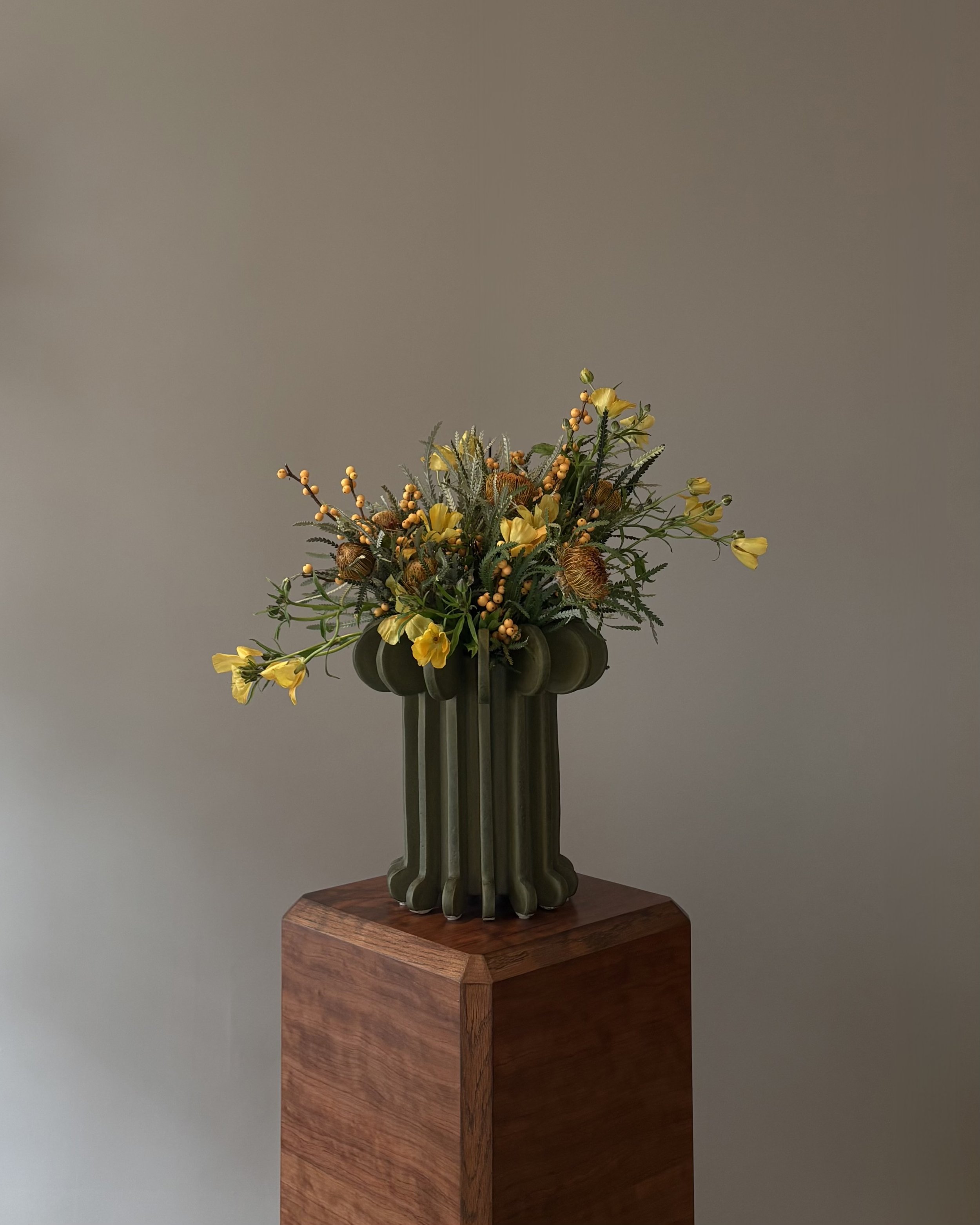 A floral arrangement with yellow flowers and greenery in a unique, deep green vase on a wooden pedestal against a plain wall.