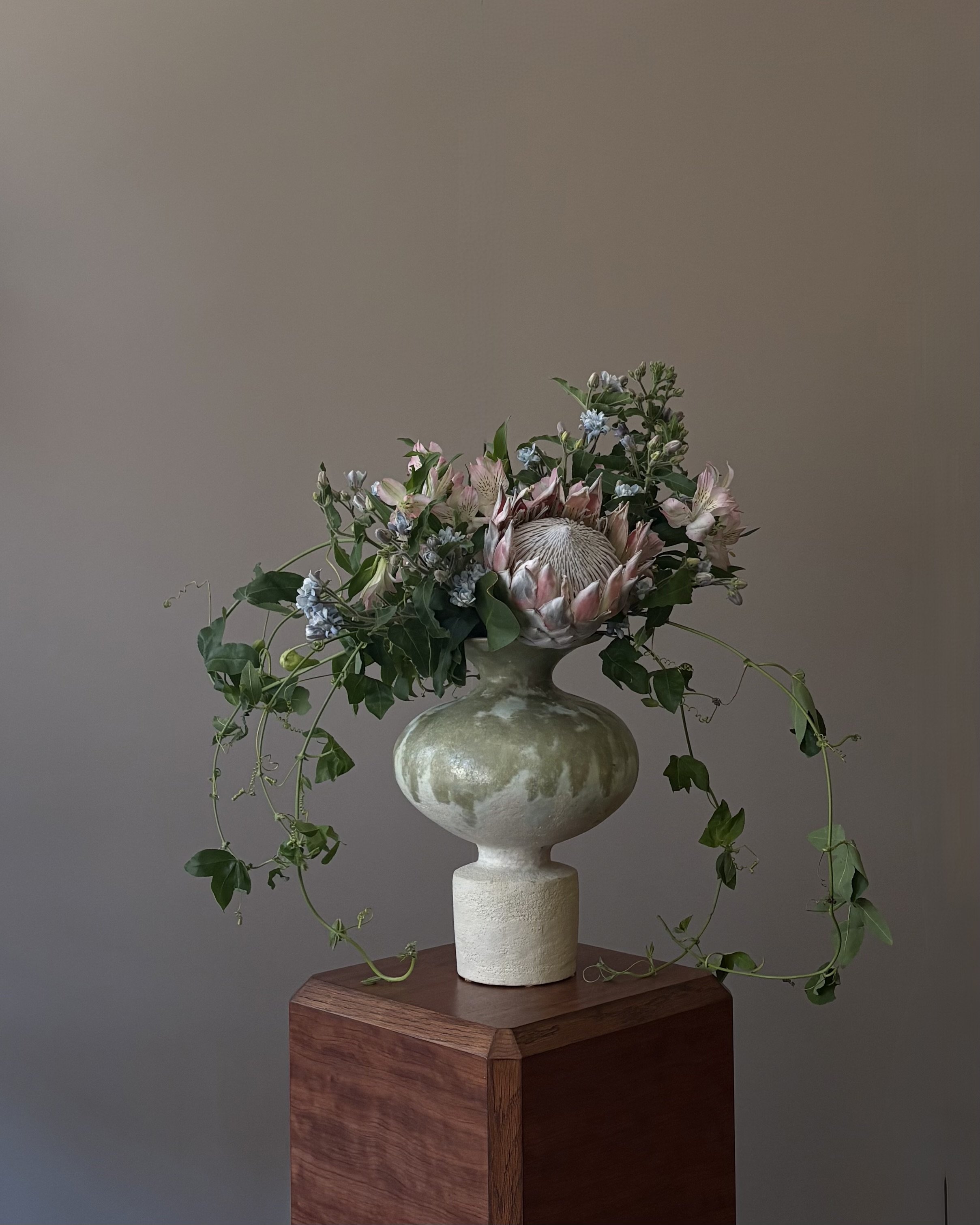 A ceramic vase with a large protea flower and assorted greenery, placed on a wooden pedestal against a plain neutral background.