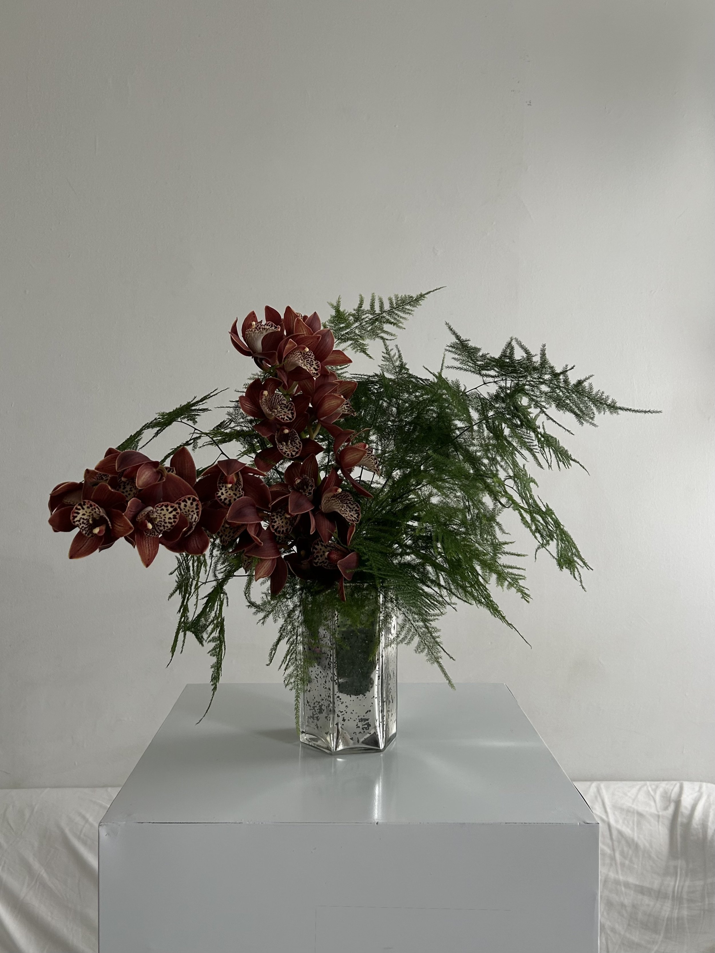 A floral arrangement with red orchids and green foliage in a clear glass vase on a white table against a white wall.