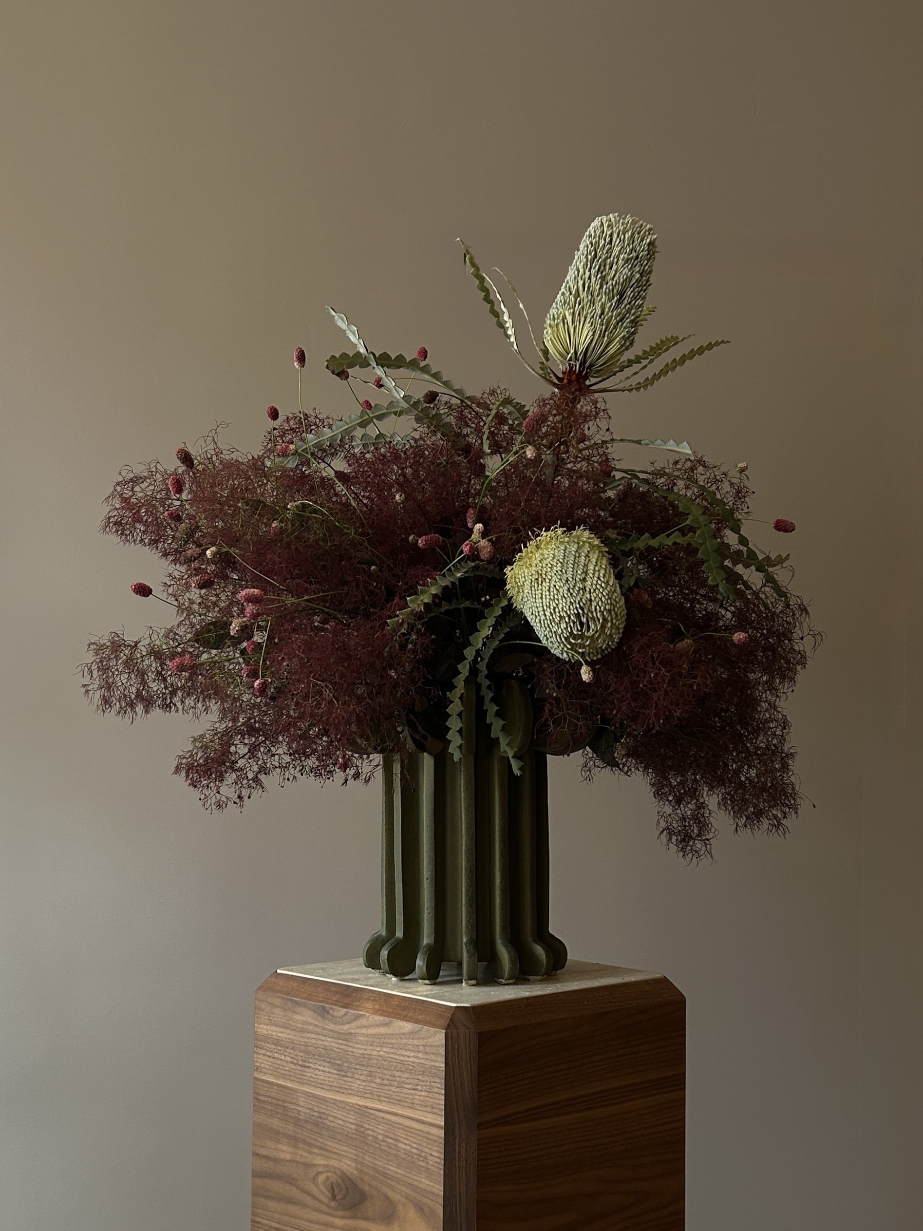 A floral arrangement in a green vase with large green and white protea flowers, surrounded by reddish-brown foliage and small pink buds, placed on a wooden pedestal.