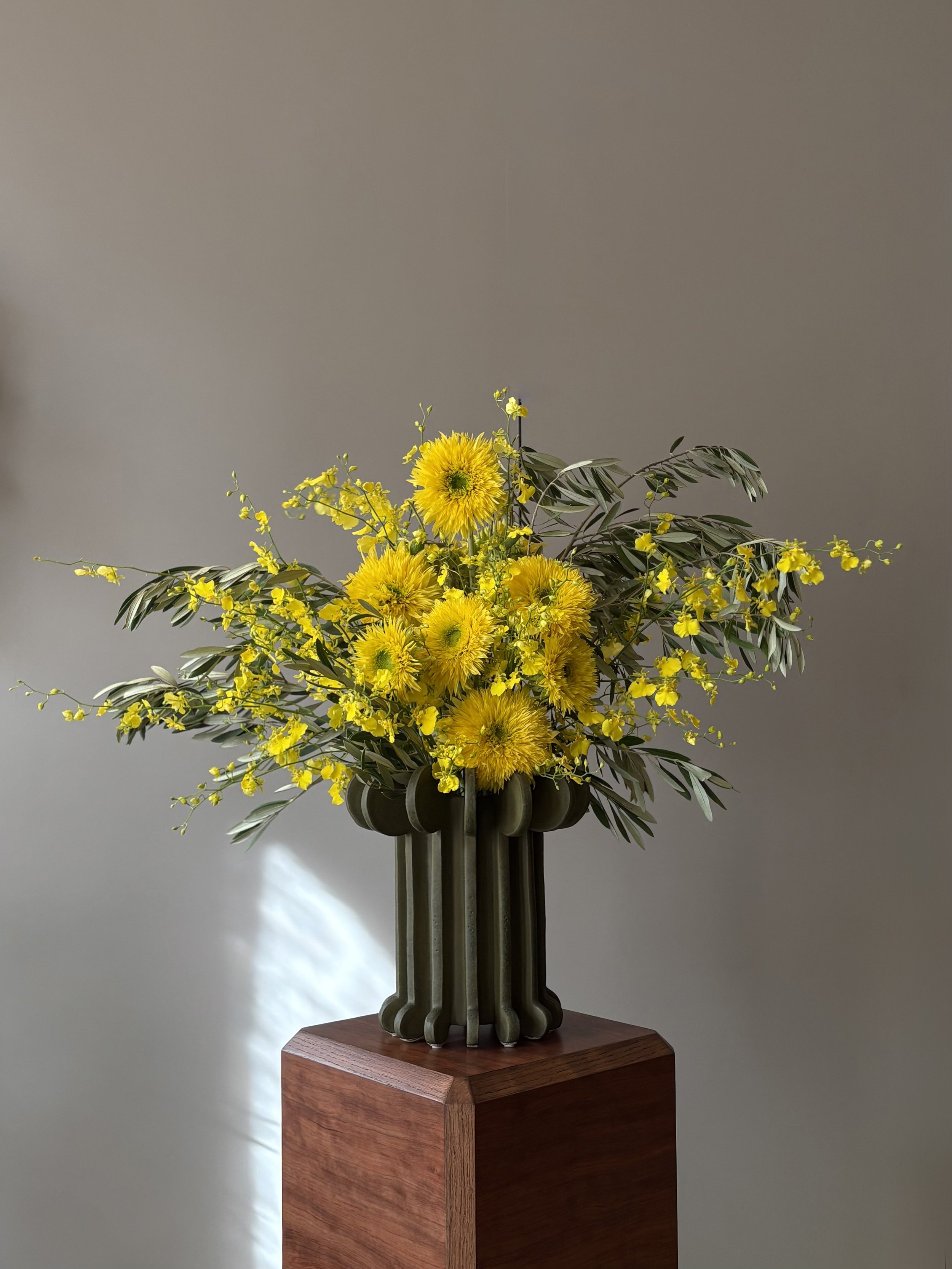 A vase with a yellow flower arrangement on a wooden pedestal against a gray wall.