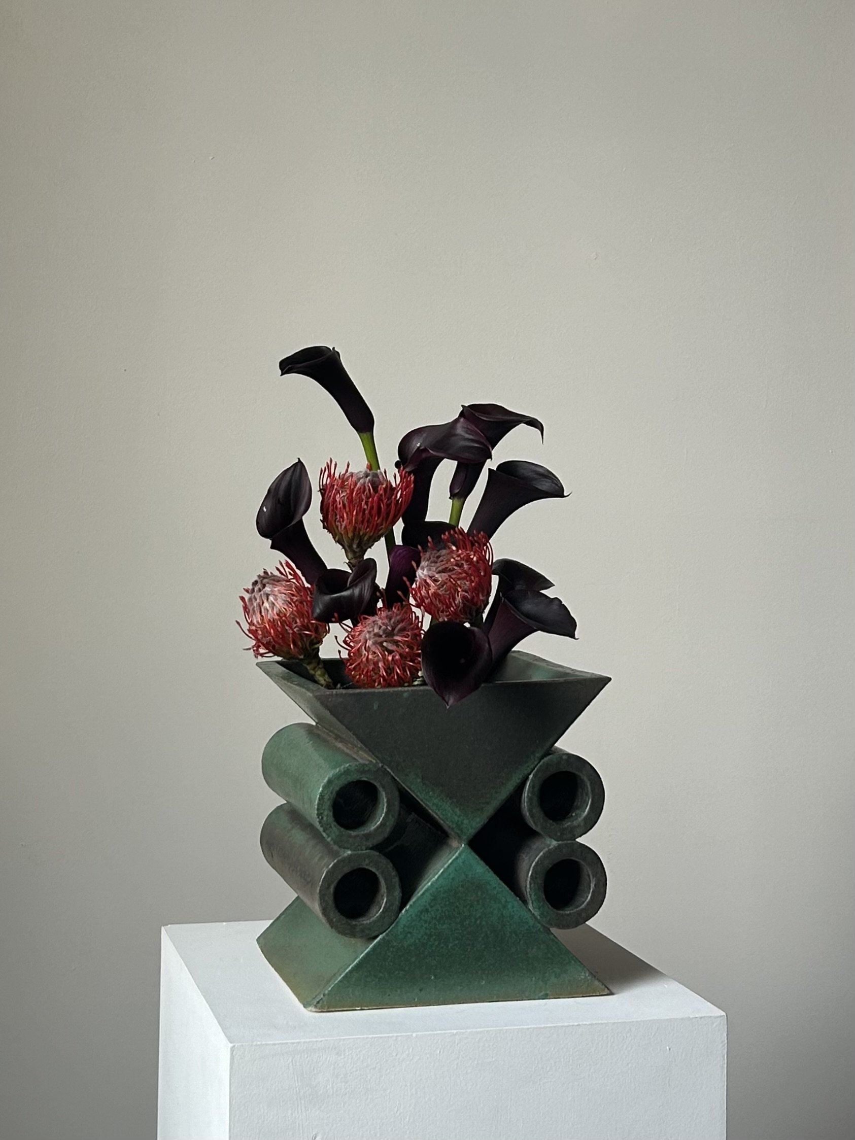 A modern sculpture with geometric metallic shapes, holding a floral arrangement of dark purple calla lilies and red protea flowers, placed on a white pedestal against a plain light-colored wall.