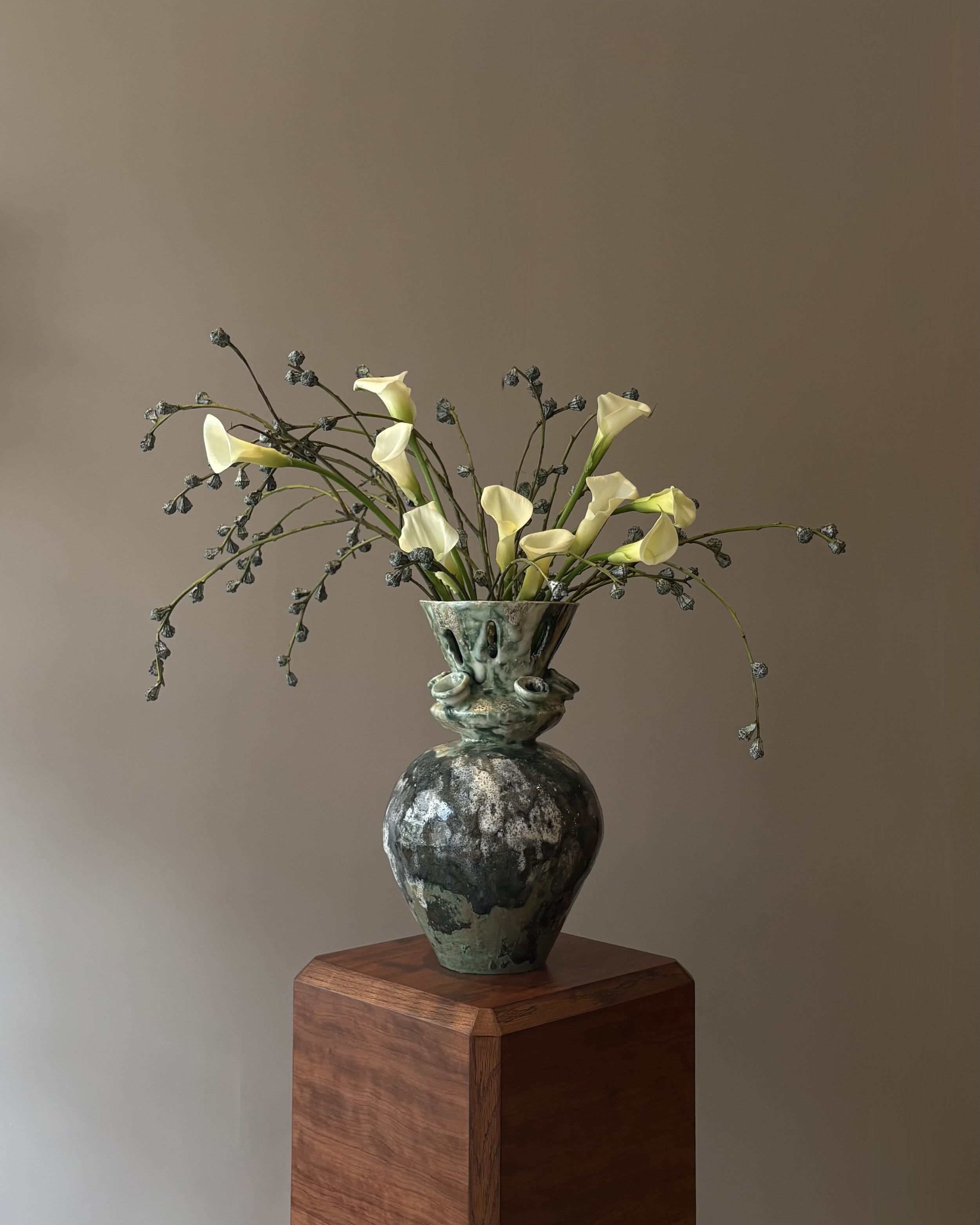 A ceramic vase with a face design, holding white calla lilies and dark, thin, curving branches with small buds, sits on a wooden pedestal against a plain wall.