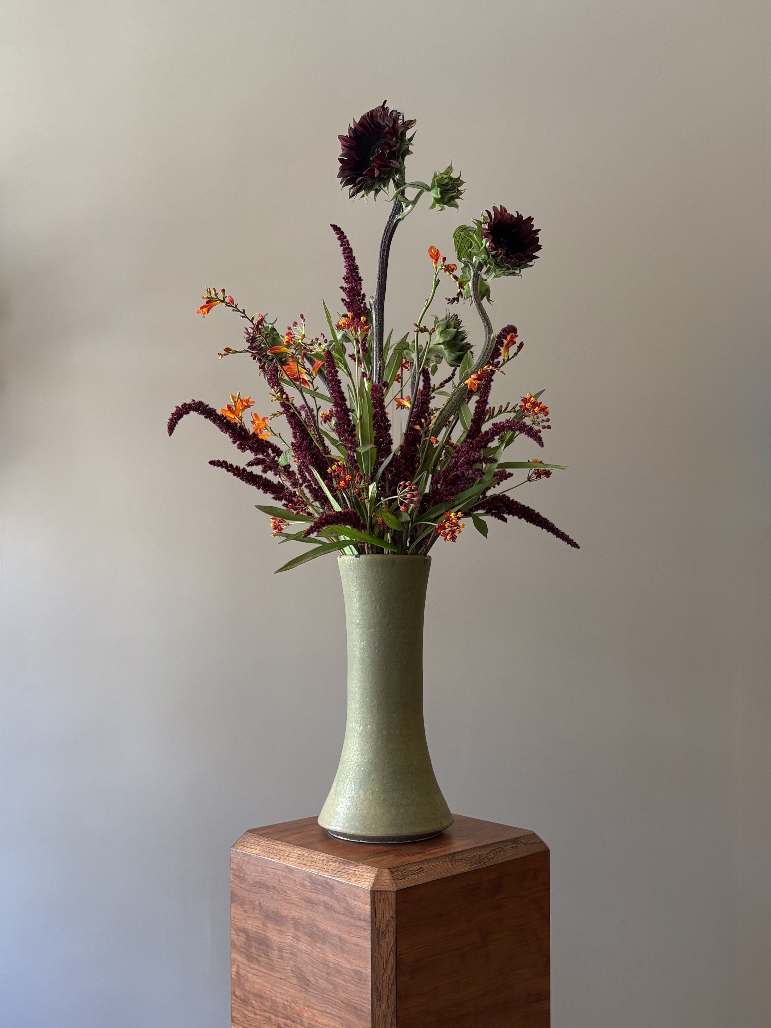 A tall green ceramic vase with a narrow opening on a wooden pedestal, holding a mixed flower arrangement with dark purple and reddish flowers and green foliage.