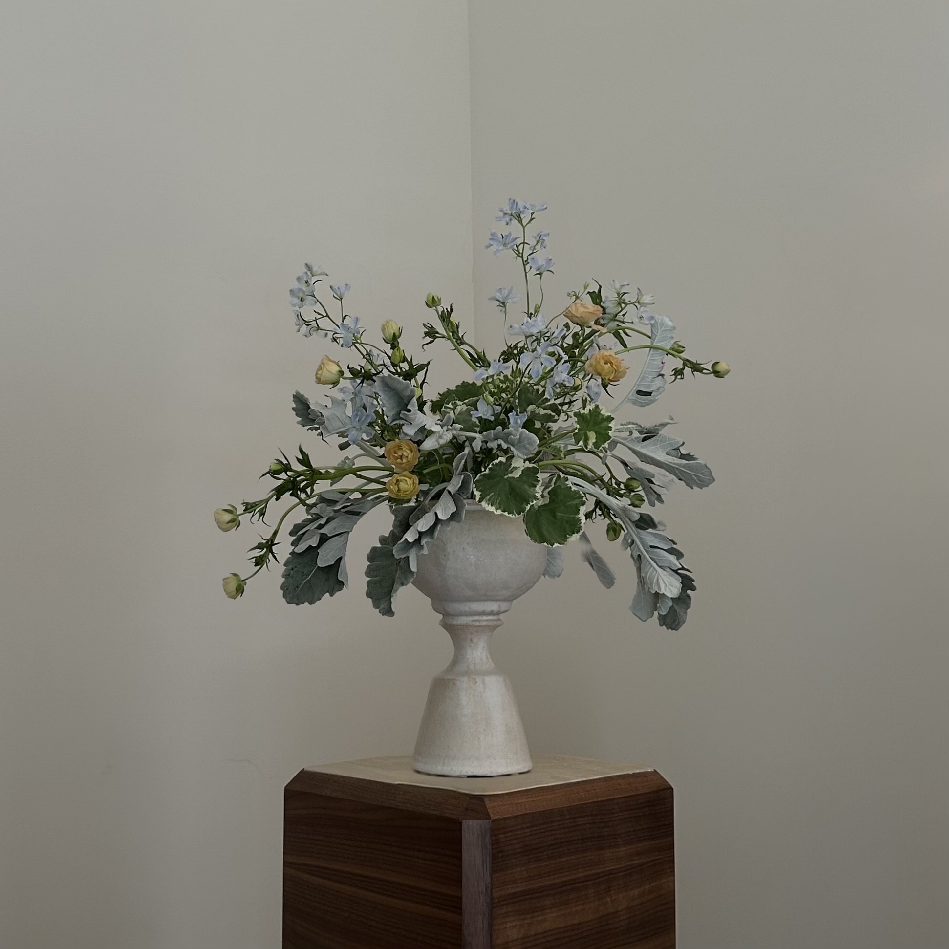 A floral arrangement in a white vase with leaves and small flowers on a wooden stand against a plain wall.
