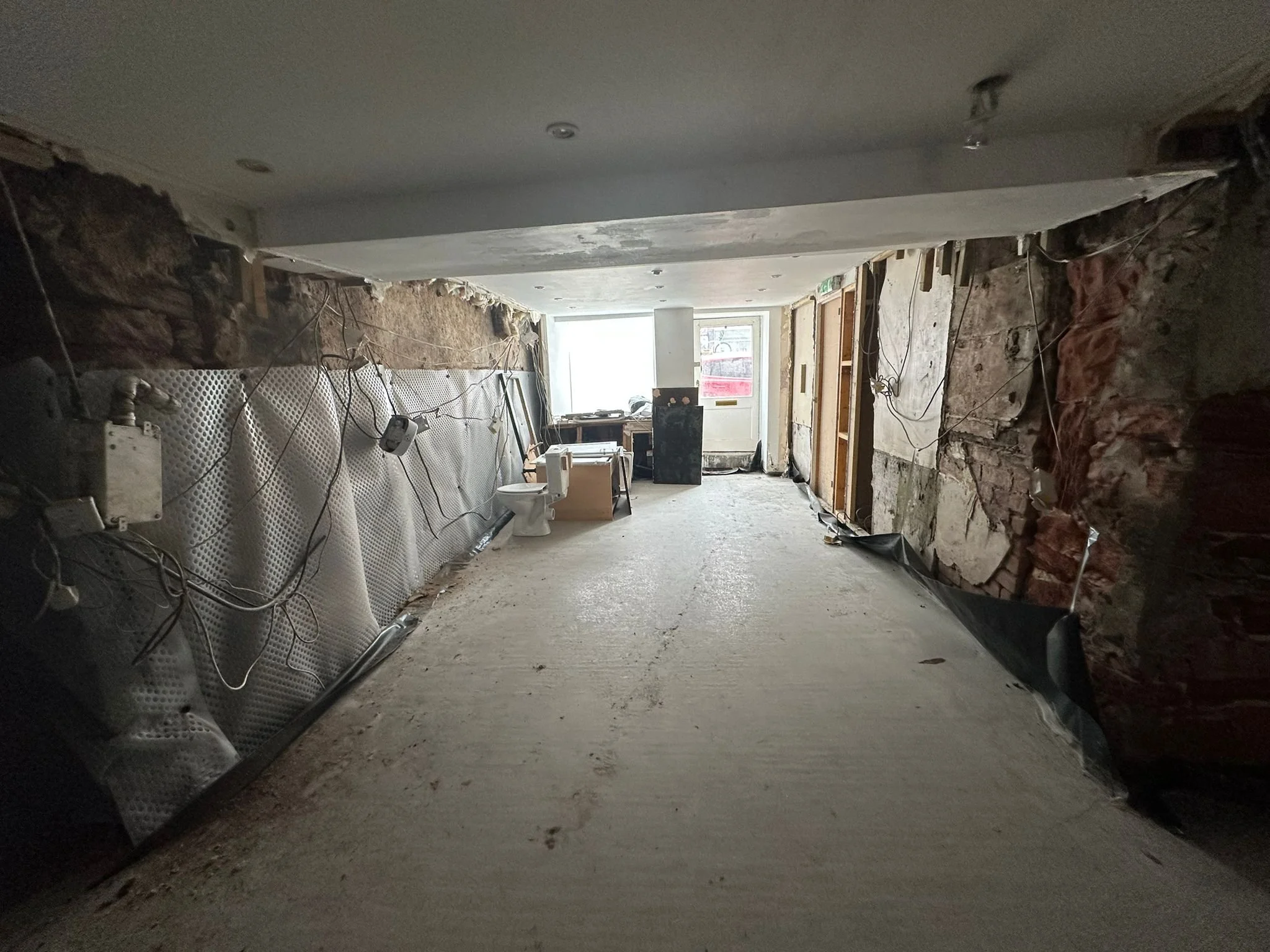 Interior of an under-construction or renovated building, showing exposed walls with insulation and wiring, a partially finished floor, a toilet, a desk, and a door leading outside.