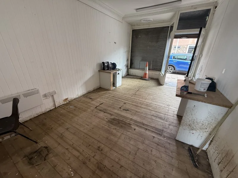 Empty room with worn wooden floor, white paneled walls, a small white cabinet, a black backpack, and a chair. Large window with partially closed blinds, and a traffic cone outside. Small countertop with cleaning supplies inside.