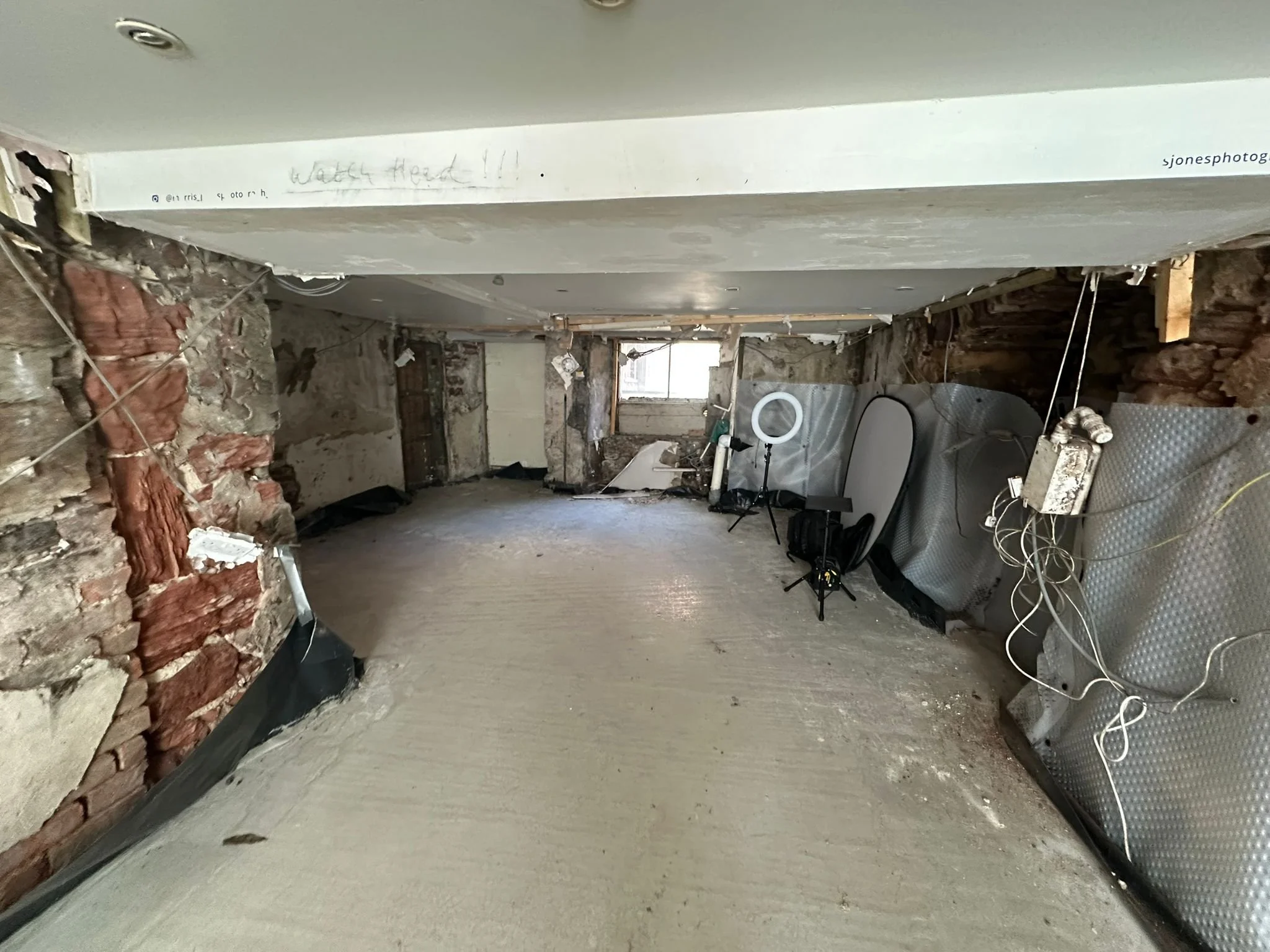 A room under renovation with exposed brick walls, wires hanging, a ring light, a monitor, and some equipment.