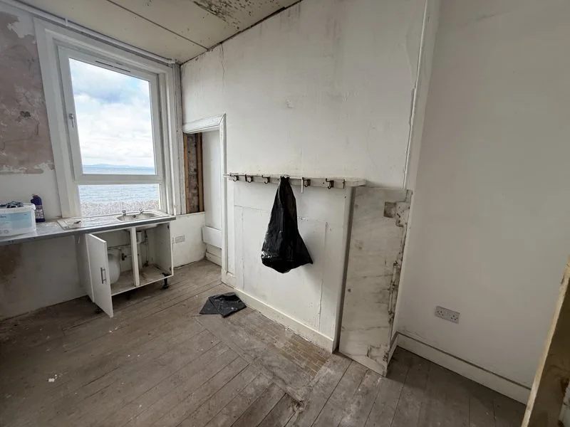 Empty room with a large window showing a view of the sea, an open sink cabinet, a black plastic bag hanging on a hook, and a worn wooden floor.