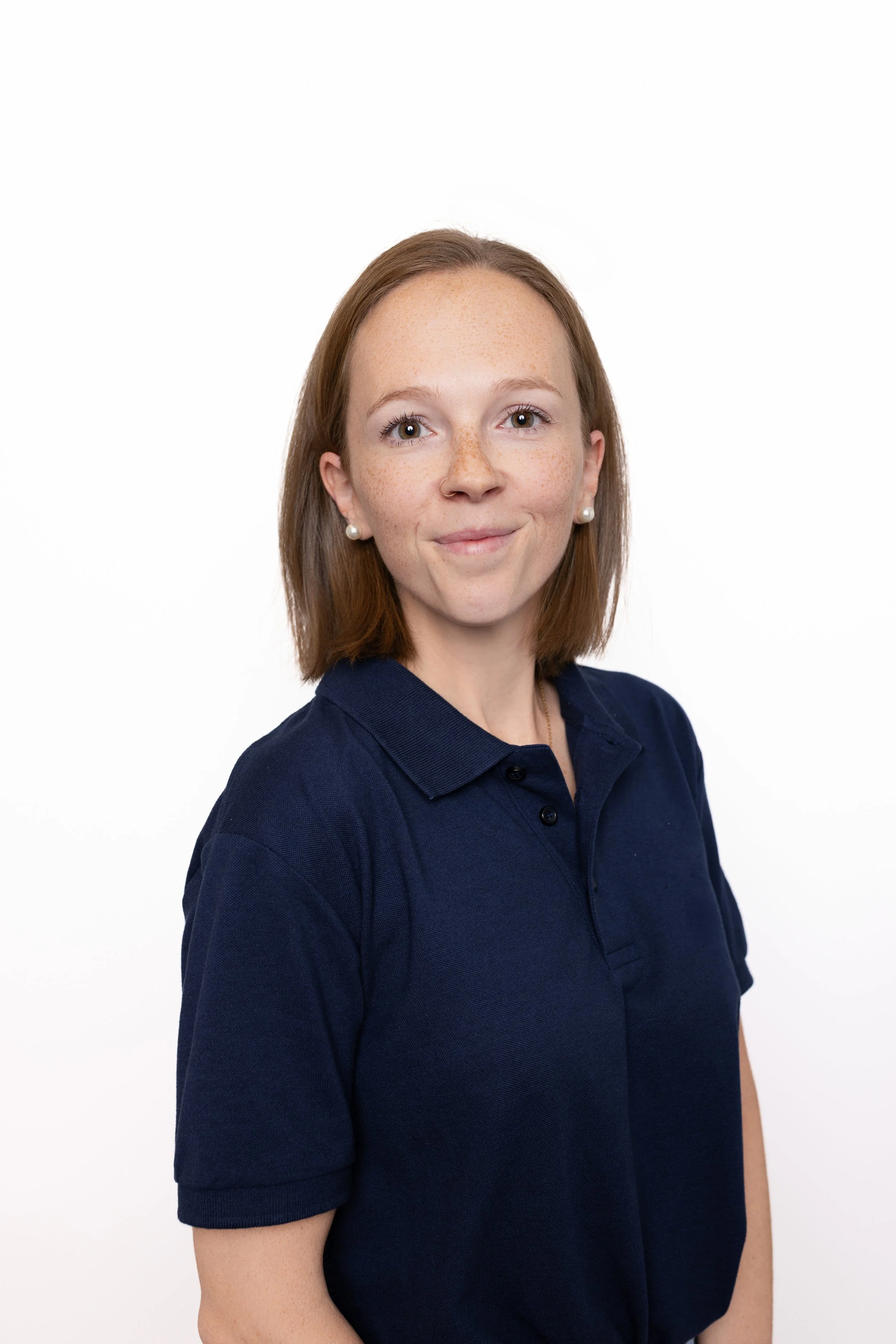 Porträt einer jungen Frau mit roten Haaren, Piercings, Pearls, in einer dunkelblauen Polo-Shirt vor weißem Hintergrund.