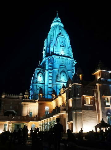 Night view of a brightly lit blue temple or palace with a tall spire, surrounded by buildings and people in the foreground.