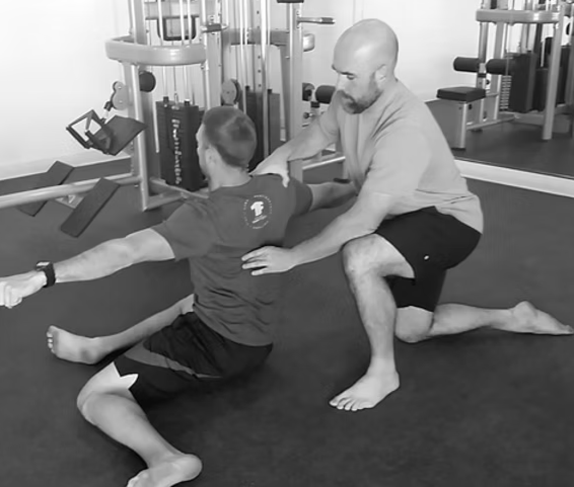 A man assisting a young boy with a stretching or exercise stretch in a gym, with exercise equipment in the background.