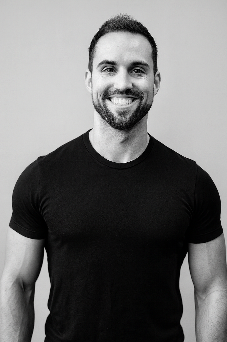 A smiling man with a beard and short hair, wearing a black t-shirt, in front of a plain light-colored background.