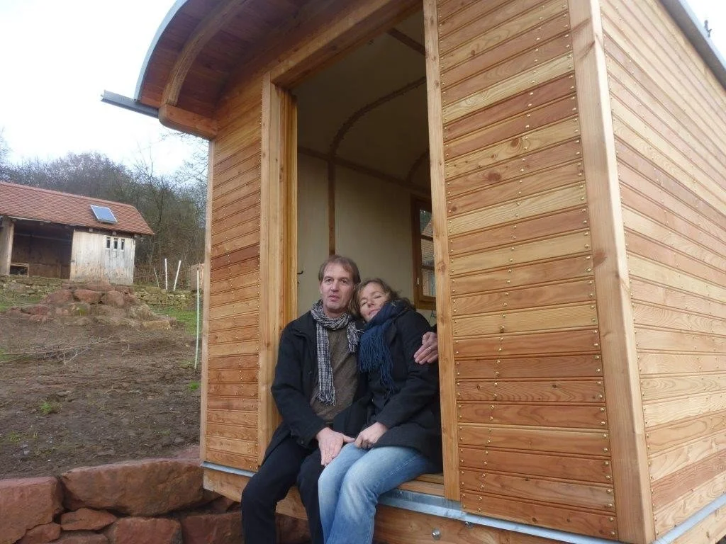Paar sitzt in einer kleinen Holzhütte, die im Bau ist, auf einem Fensterbrett. Hintergrund zeigt Baumlandschaft und eine kleinere Holzhütte.