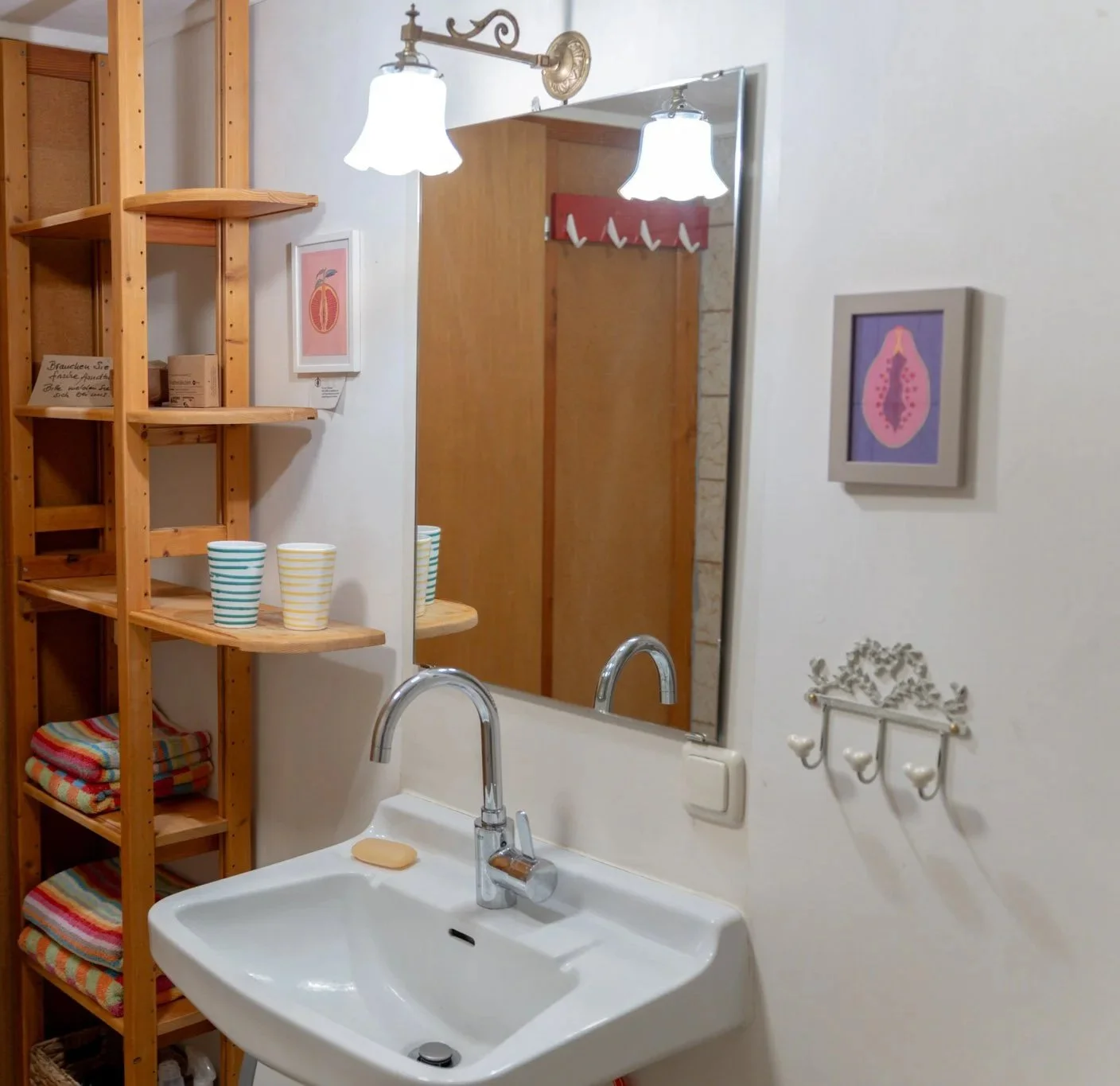 Ein Badezimmer mit Waschbecken, Spiegel, Holzregal mit bunten Handtüchern und Bechern, Wanddekorationen, einem Lichtstrahler und einem Handtuchhalter