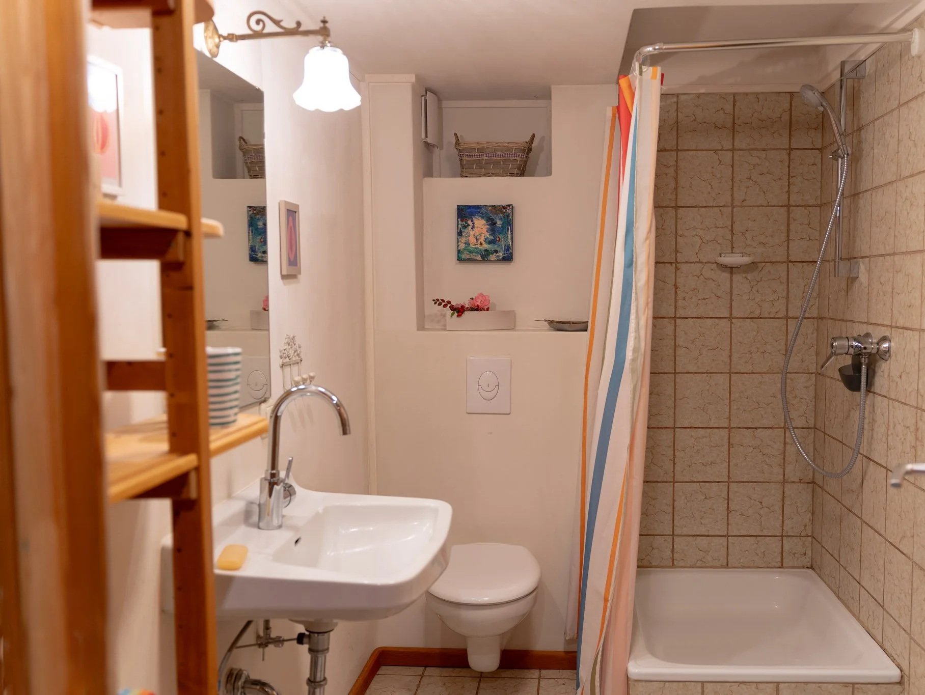 Kleines Badezimmer mit Waschbecken, Toilette und Dusche, aus Holzregalen, Bilder und dekorative Gegenstände auf den Regalen, bunte Duschvorhang.