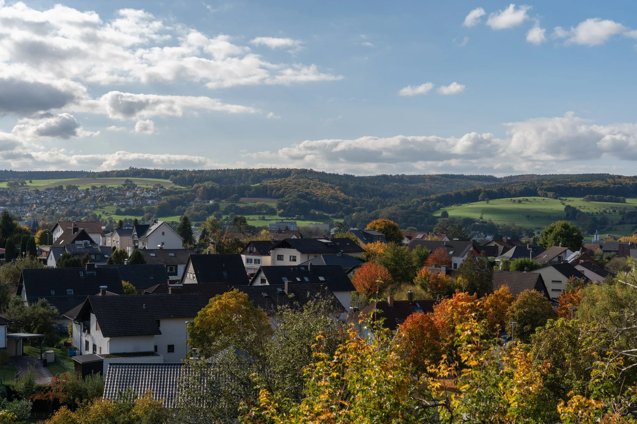 Blick auf ein Dorf mit Hausdächern und bunten Herbstbäumen vor Hügeln und einer bewölkten Himmelslandschaft.
