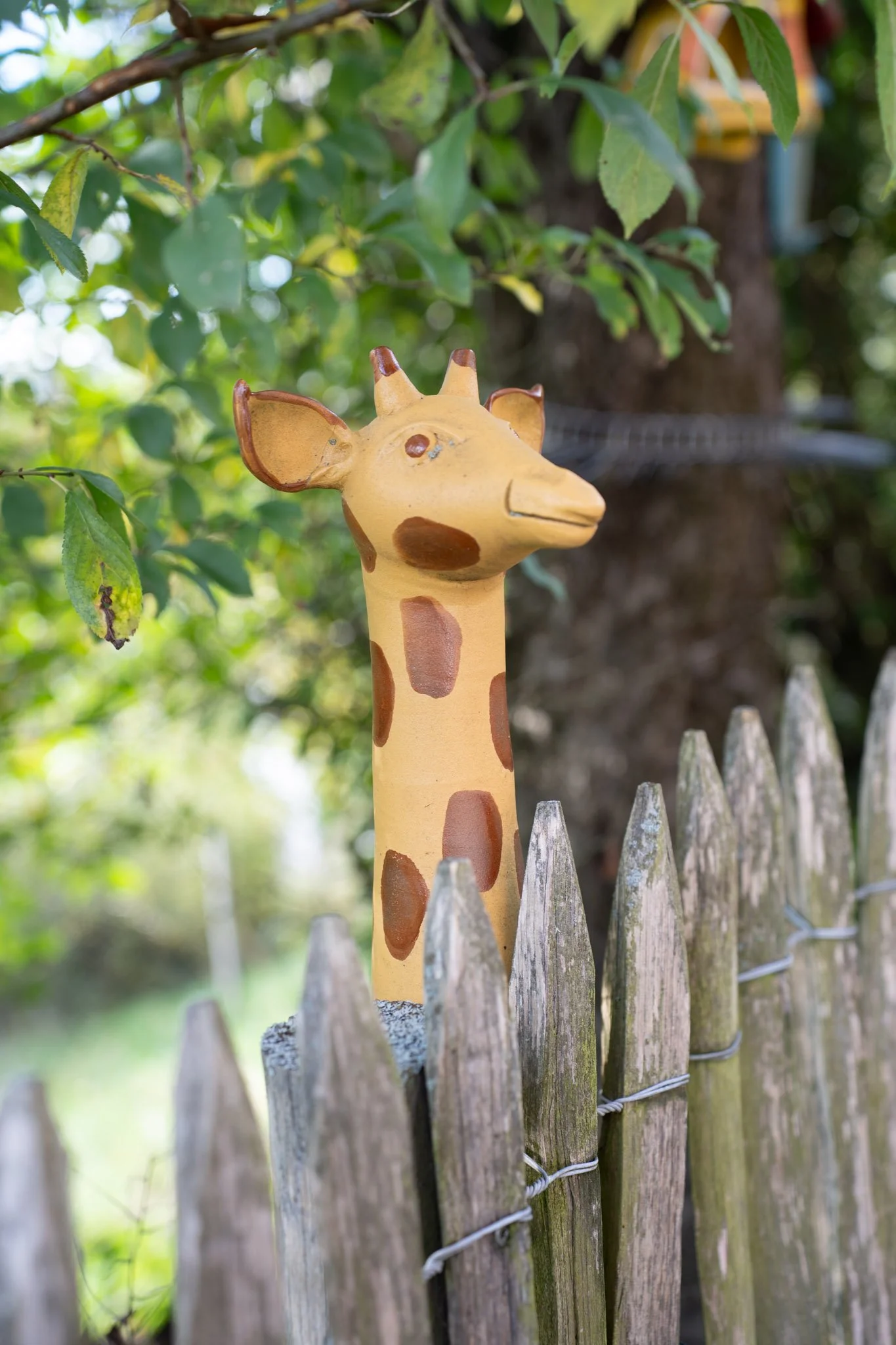 Ein bemalter Zementstorch in Form eines Giraffenkopfes, der über einem Holzzaun steht, vor grünen Blättern.