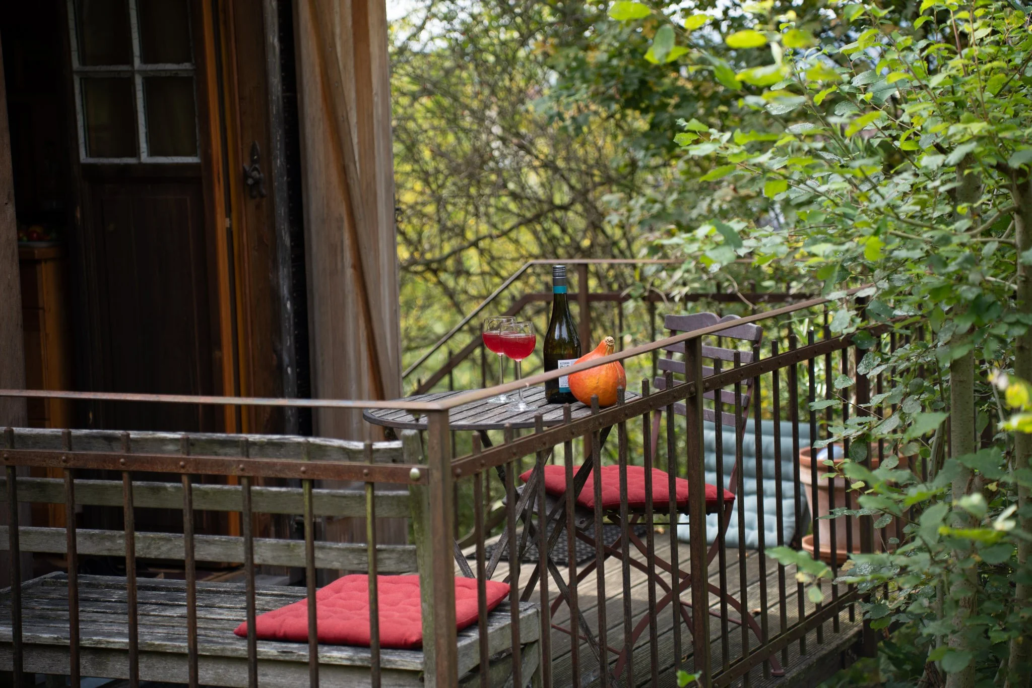 Ein kleiner Balkon mit einem runden Tisch, auf dem eine Flasche Wein, ein Weinglas mit Rotwein und eine orange Vase stehen. Es gibt zwei Stühle mit roten Sitzkissen, um den Tisch herum, umgeben von Bäumen und grüner Natur.