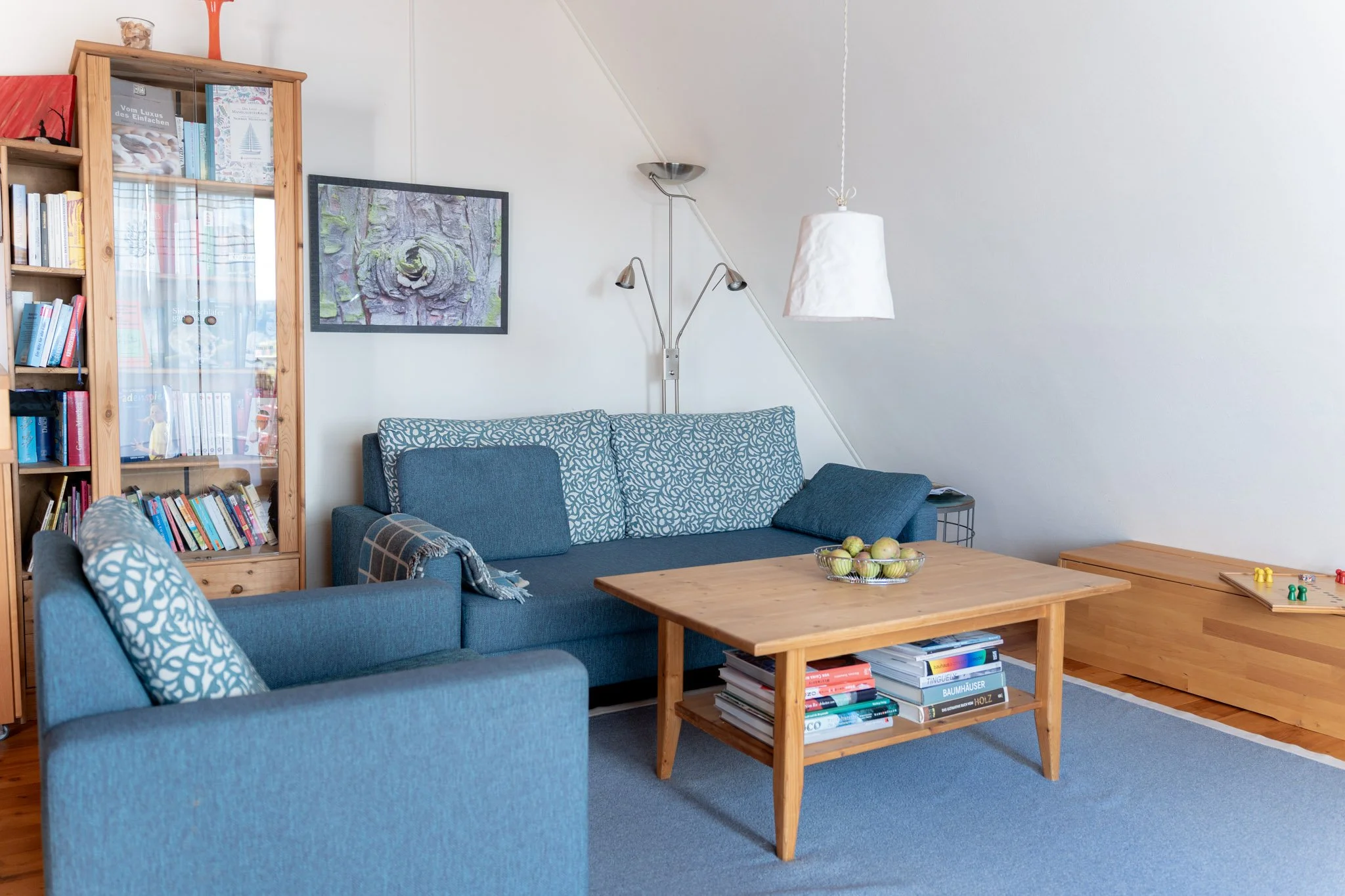 Gemütliches Wohnzimmer mit blauer Couch, einem Holztisch, Bücherregal, einer Bodenlampe, hängender Lampenschirm und einem Spielbrett auf dem Boden