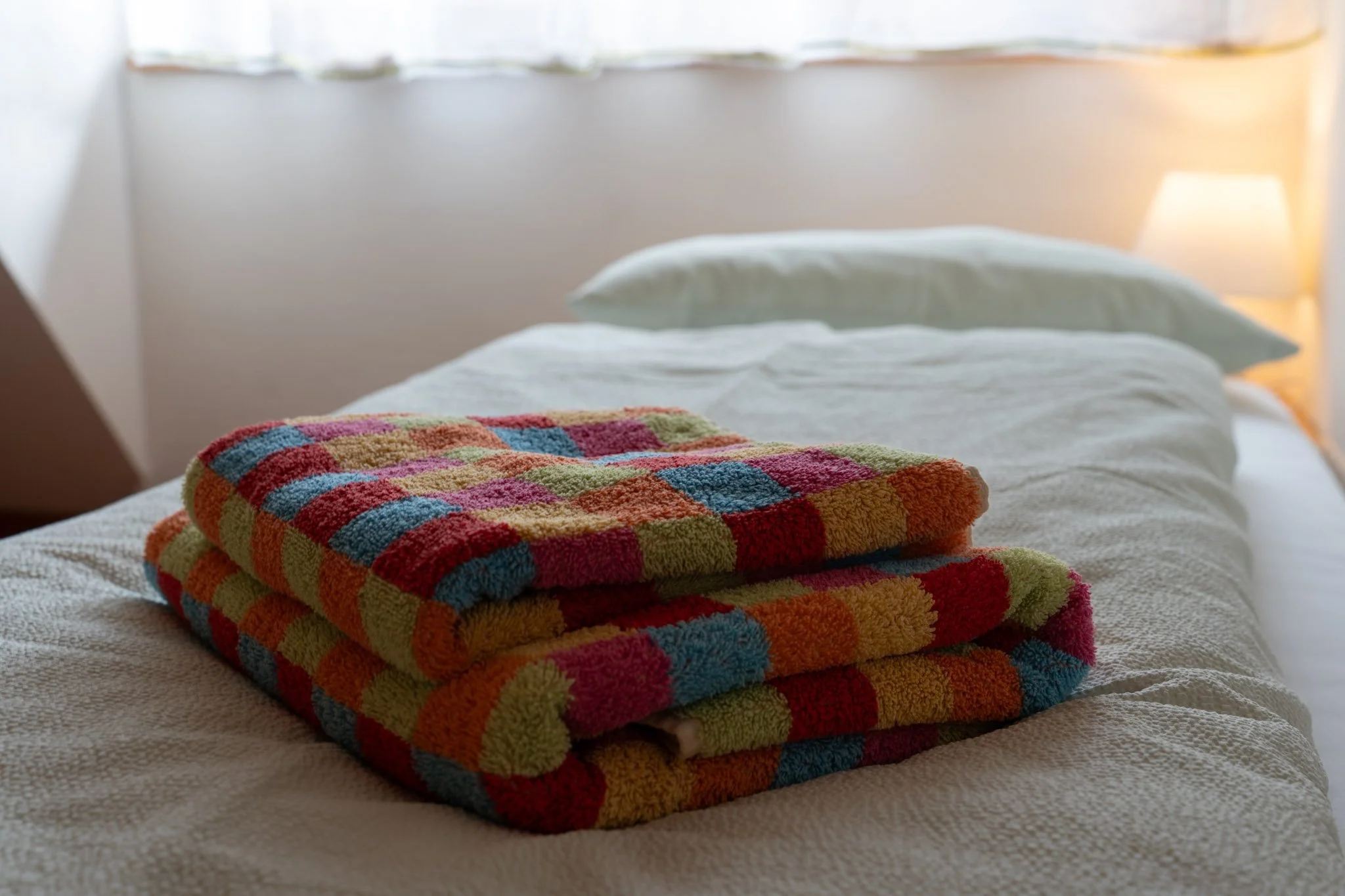 Bett mit weißen Bettdecken und Kissen, davor gefaltete bunte Handtücher in bunten Karomustern.