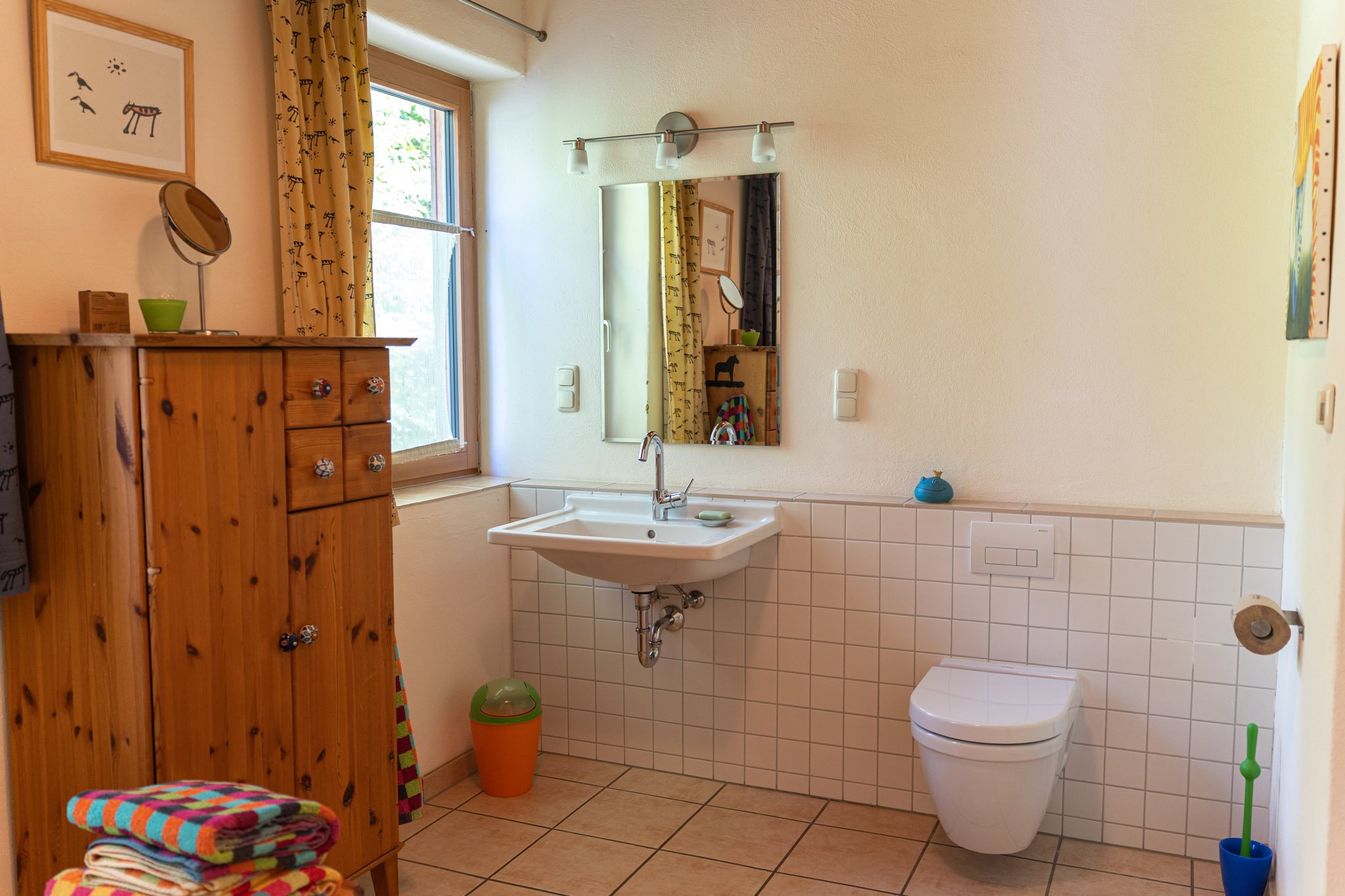 Ein Badezimmer mit einer weißen Wand, einem quadratischen Wandspiegel, einem Fenster mit gelben Vorhängen, einer weißen Toilette, einem weißen Waschbecken und einer Holzmöbel auf der linken Seite. Es gibt bunte Handtücher auf einem Regal im Vordergru