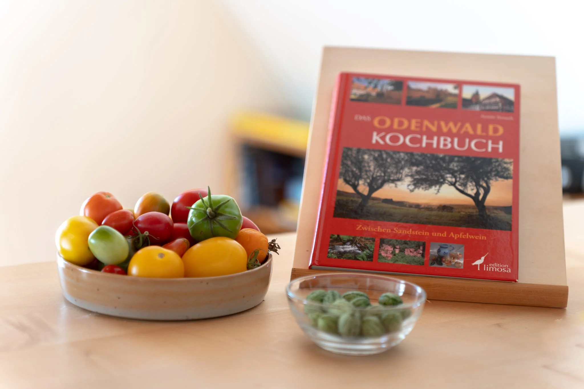 Bunte Tomatenschale, ein Kochbuch über den Odenwald, und eine kleine Schüssel mit grünen Bohnen auf einem Holztisch.