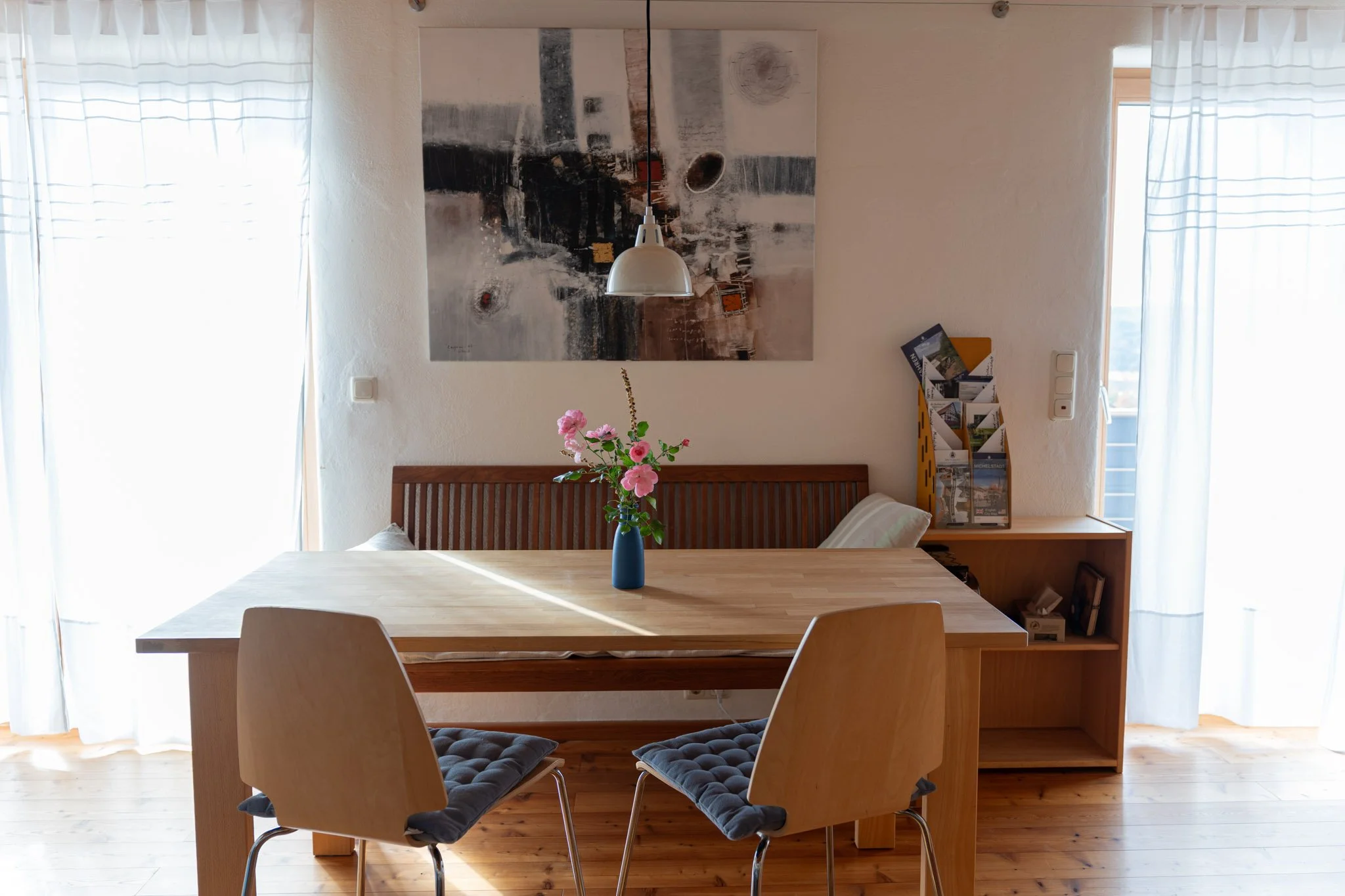 Ein Esszimmer mit einem großen Holztisch, zwei Stühlen mit gepolsterten Sitzen, einer blauen Vase mit rosa Blumen, einem Wandbild, einem Regal mit Broschüren, viel natürlichem Licht durch Fenster mit weißen Vorhängen.