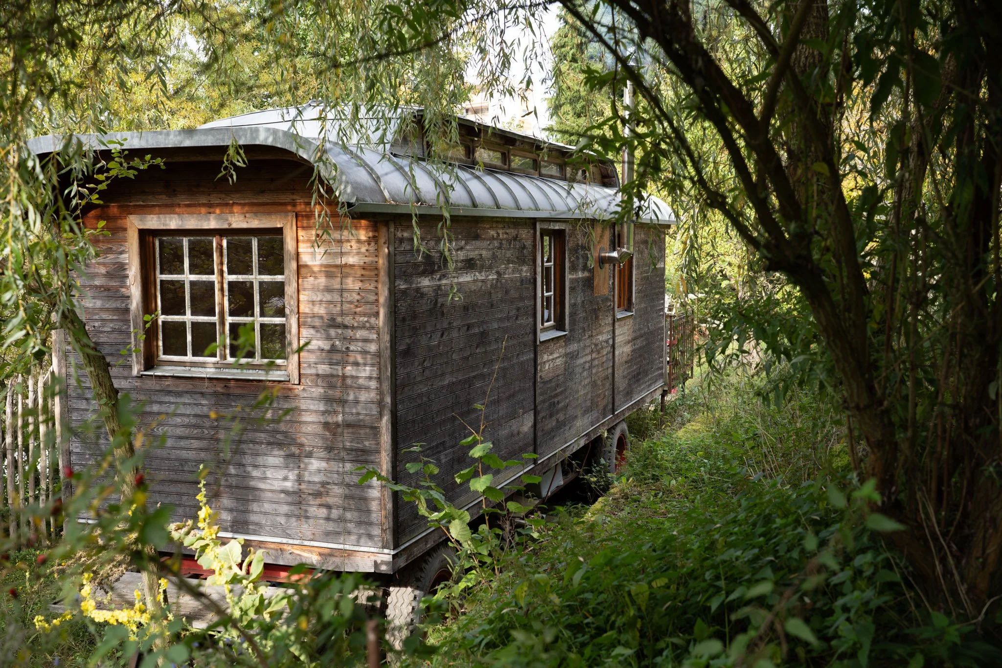 Ein altes, fahrbares Holzhaus mit mehreren Fenstern steht in einem Garten, umgeben von Büschen und Bäumen.
