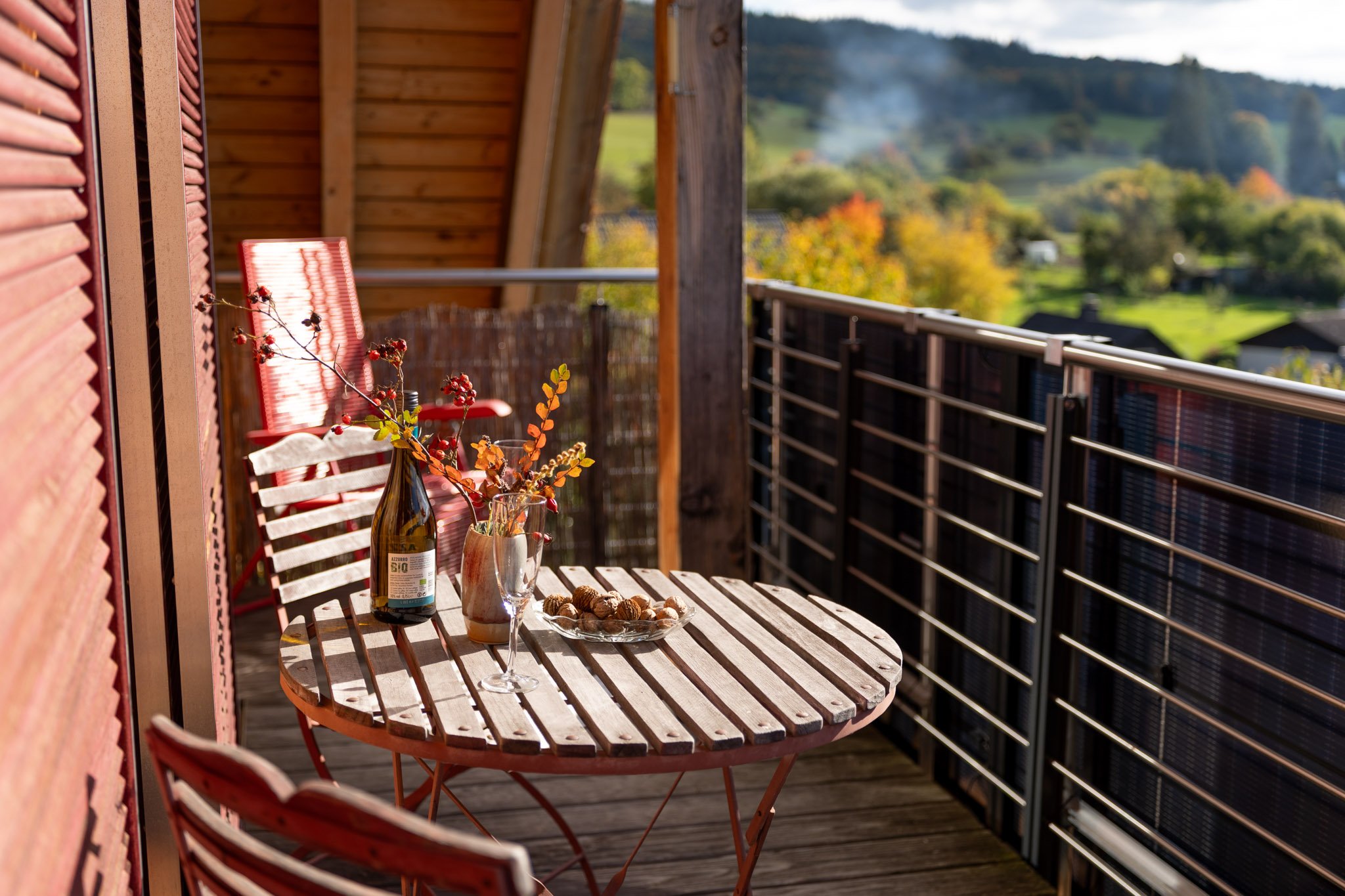 Balkon mit Holz Tisch und Stühlen, Dekoration mit Blumen und Wein, Blick auf grüne Hügel und Bäume im Herbst, sonniges Wetter.