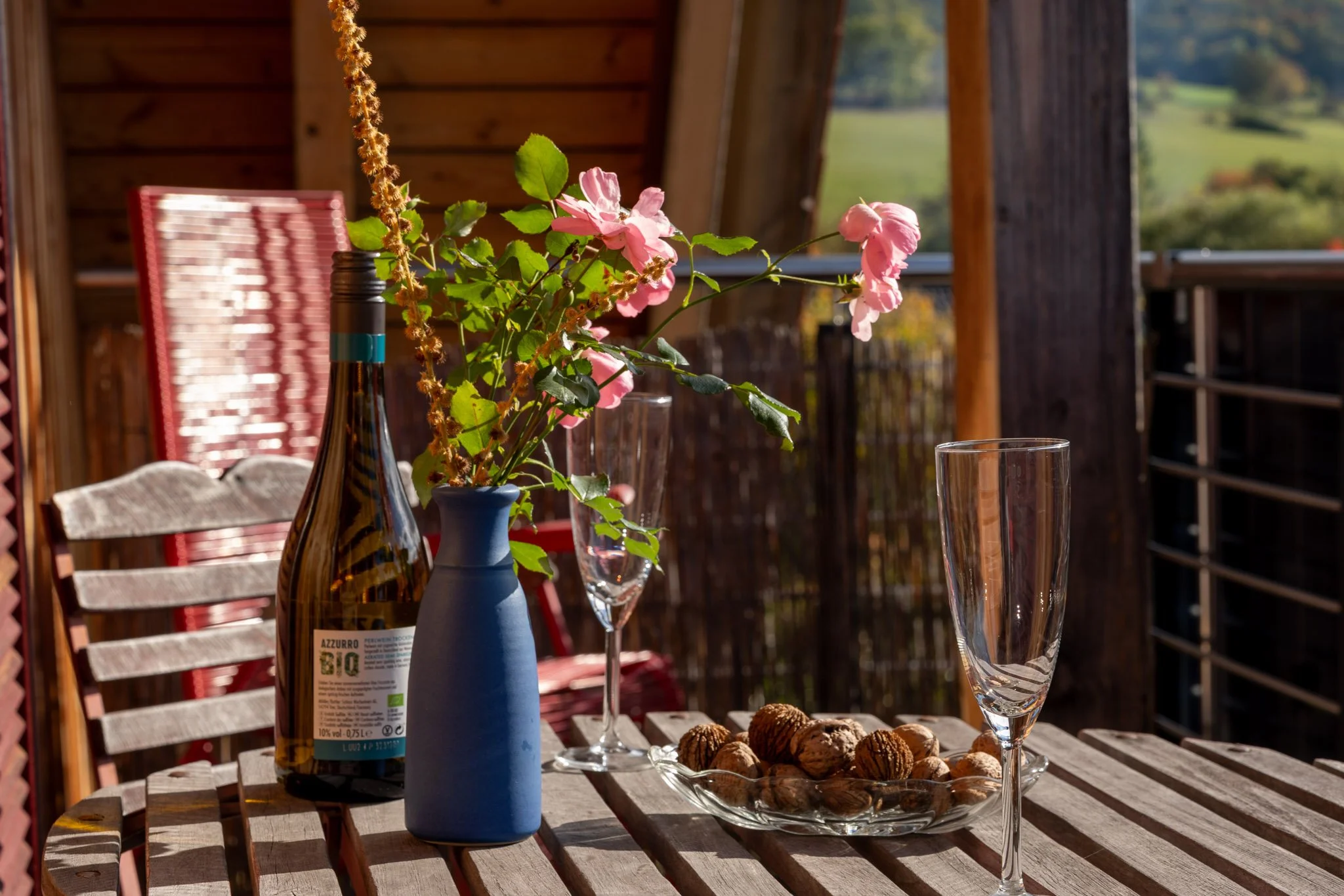 Ein rustikaler Holztisch auf einer Balkonveranda mit einer blauen Vase, pinken Blumen, leeren Weingläser, einer Weinflasche und einer Schale mit Walnüssen bei Sonnenlicht, im Hintergrund grünes Hügelland.