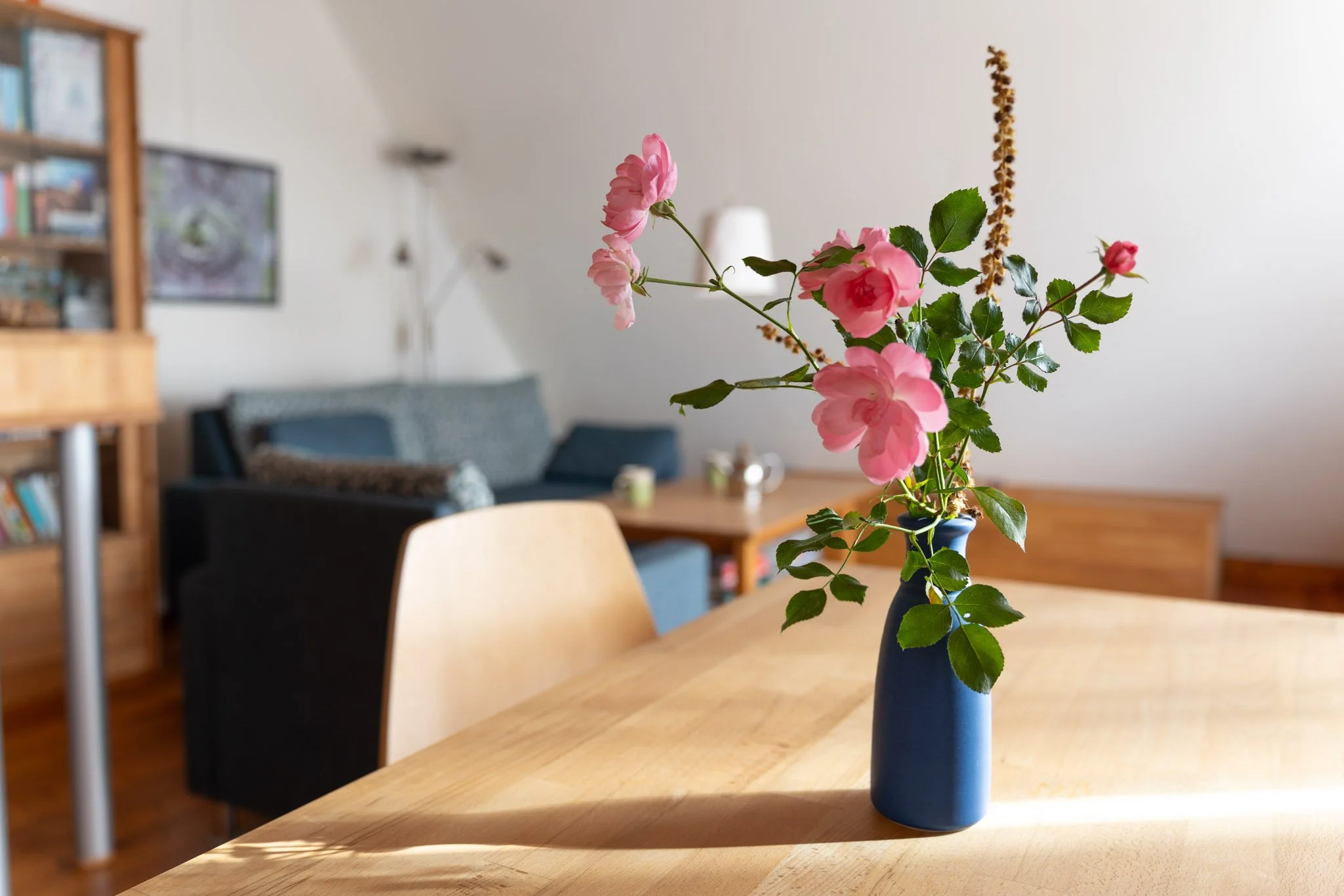 Ein blauer Vase mit rosa Blumen auf einem Holztisch im Wohnzimmer, im Hintergrund ein Sofa und Bücherregal.