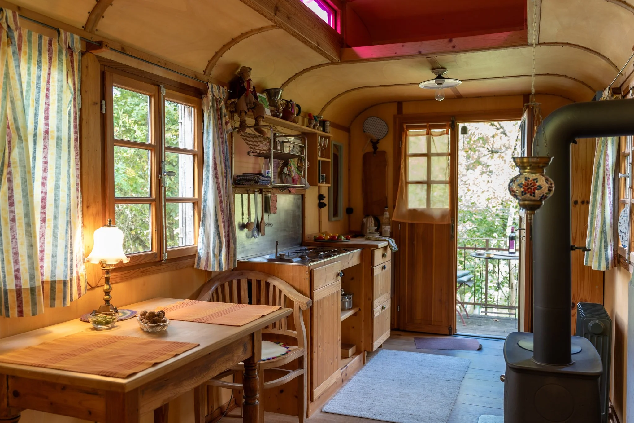 Gemütliche Küche mit Holzinterieur, Fenster mit striped Gardinen, Esstisch mit Tischläufern, kleine Küchecke mit Gasherd, Türen zum Balkon mit Holzfenster und viele dekorative Gegenstände.