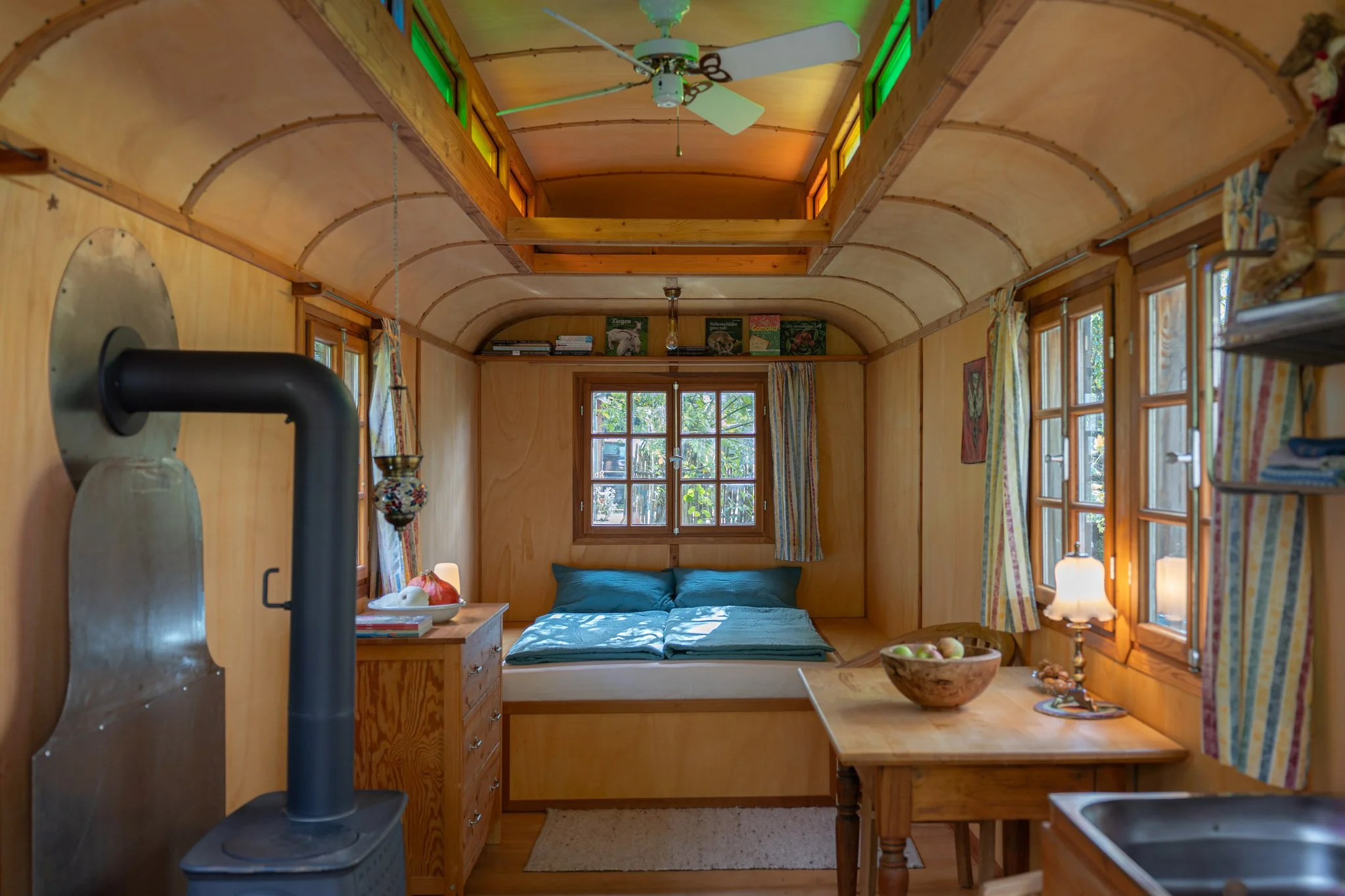 Innenraum eines kleinen, holzgetäfelten Hauses mit mehreren Fenstern, einem Bett vor der Wand, einer Lampe auf einem Nachttisch, einem Holztisch mit einem Obstkorb, einer Küchenzeile mit Mikrowelle und einem Holzofen, bunter Deckenleiste, Holzkasten 