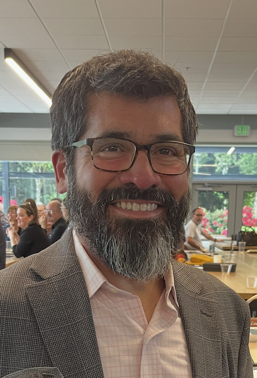 A man with glasses and a beard smiling in an indoor setting with people and large windows in the background.