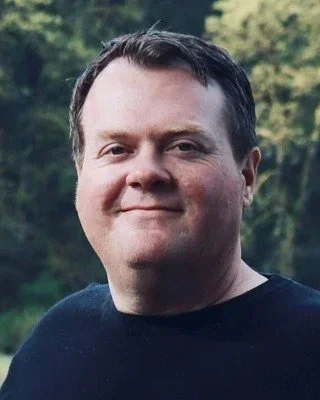 A man with short dark hair smiling outdoors, with trees in the background.
