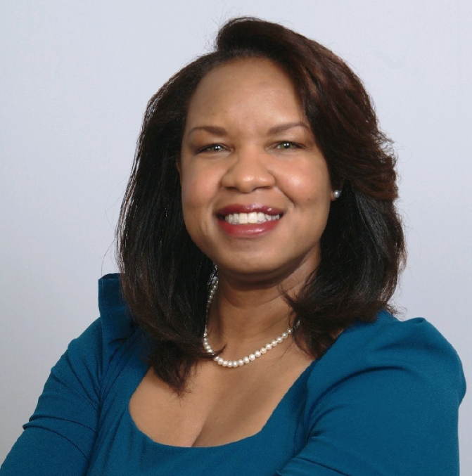 A smiling African American woman with shoulder-length dark hair, wearing a blue dress, pearl necklace, and pearl earrings, posed against a plain light background.